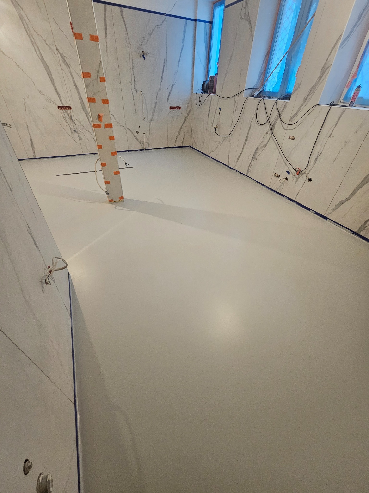 Jasna, gładka podłoga w pomieszczeniu z marmurowymi ścianami i oknami dachowymi. Widoczne zabezpieczenia taśmą malarską i wystające kable elektryczne.