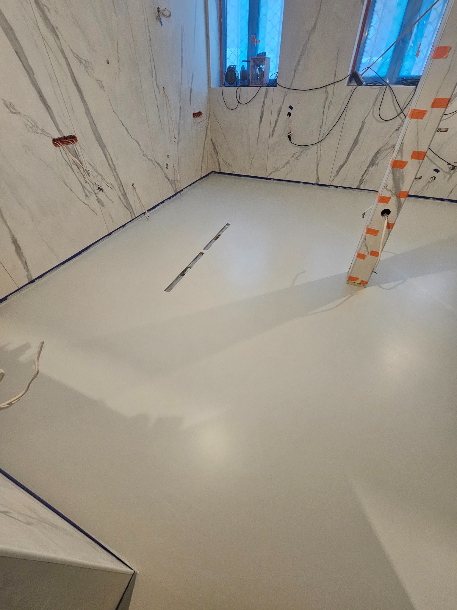 Jasna, gładka powierzchnia podłogi z odpływem liniowym, świeżo po wylaniu, zabezpieczona niebieską taśmą malarską przy ścianach wykończonych marmurem.