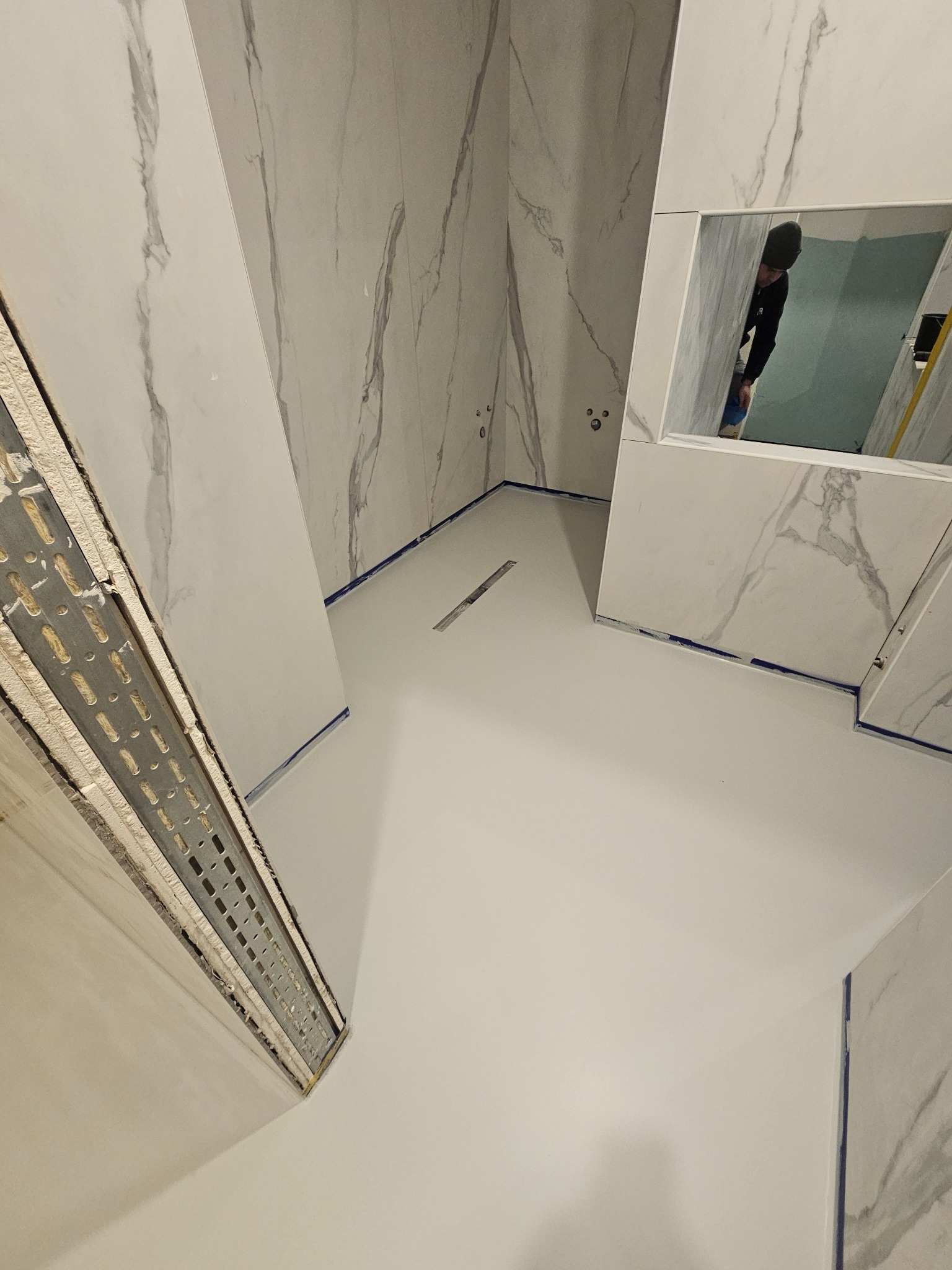 Biała, gładka posadzka w łazience z marmurowymi płytkami na ścianach. Widoczny odpływ liniowy i odbicie osoby w lustrze. Wykończenie z niebieską taśmą malarską.