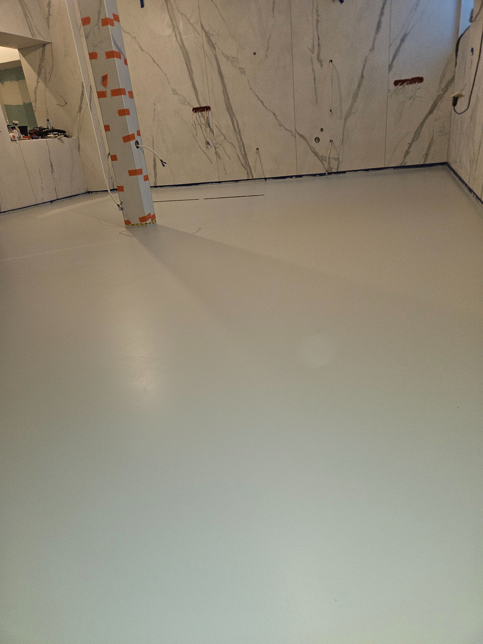 Jasnoszara, gładka podłoga w pomieszczeniu z marmurowymi ścianami. Słup zabezpieczony taśmą malarską. Widoczne przygotowania do wykończenia.