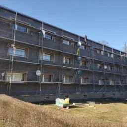 Budynek wielorodzinny w trakcie ocieplania w Strzelcach Opolskich, z widocznymi rusztowaniami i pracownikiem na wysokości. Elewacja pokryta szarą warstwą izolacyjną.