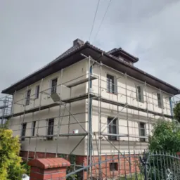 Dwupiętrowy dom w rusztowaniach podczas malowania elewacji na kremowo, z widocznym wiadrem farby. Częściowo pomalowana ściana i pracownik na rusztowaniu.