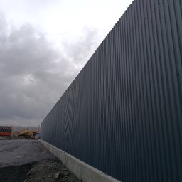 MAX - GLOBAL Maksym Kiselov - Długa, szara ściana z blachy falistej na betonowej podmurówce, widoczna z boku, z ziemią i sprzętem budowlanym w tle pod pochmurnym niebem.