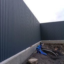 MAX - GLOBAL Maksym Kiselov - Szara, blaszana elewacja budynku z widocznymi fundamentami i instalacją w ziemi. Ujęcie z dołu, podkreślające pionowe linie blachy i surowość betonu.