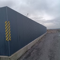 MAX - GLOBAL Maksym Kiselov - Długi, szary płot z blachy falistej na betonowej podmurówce, z żółto-czarnymi pasami ostrzegawczymi, oświetlony lampą, na tle pochmurnego nieba i hałdy ziemi.