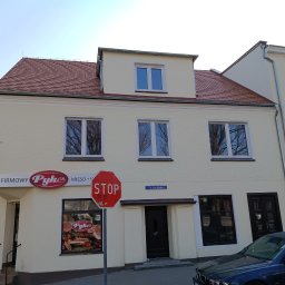 Max Global - Odnowiona elewacja budynku sklepu w Strzelcach Opolskich. Jasna fasada z czerwonym dachem, okna z odbiciami drzew. Widoczny szyld sklepu i znak STOP.