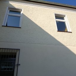 Max Global - Jasna elewacja budynku z trzema oknami, z czego jedno z kratą. Na elewacji widoczny cień, a w tle błękitne niebo.