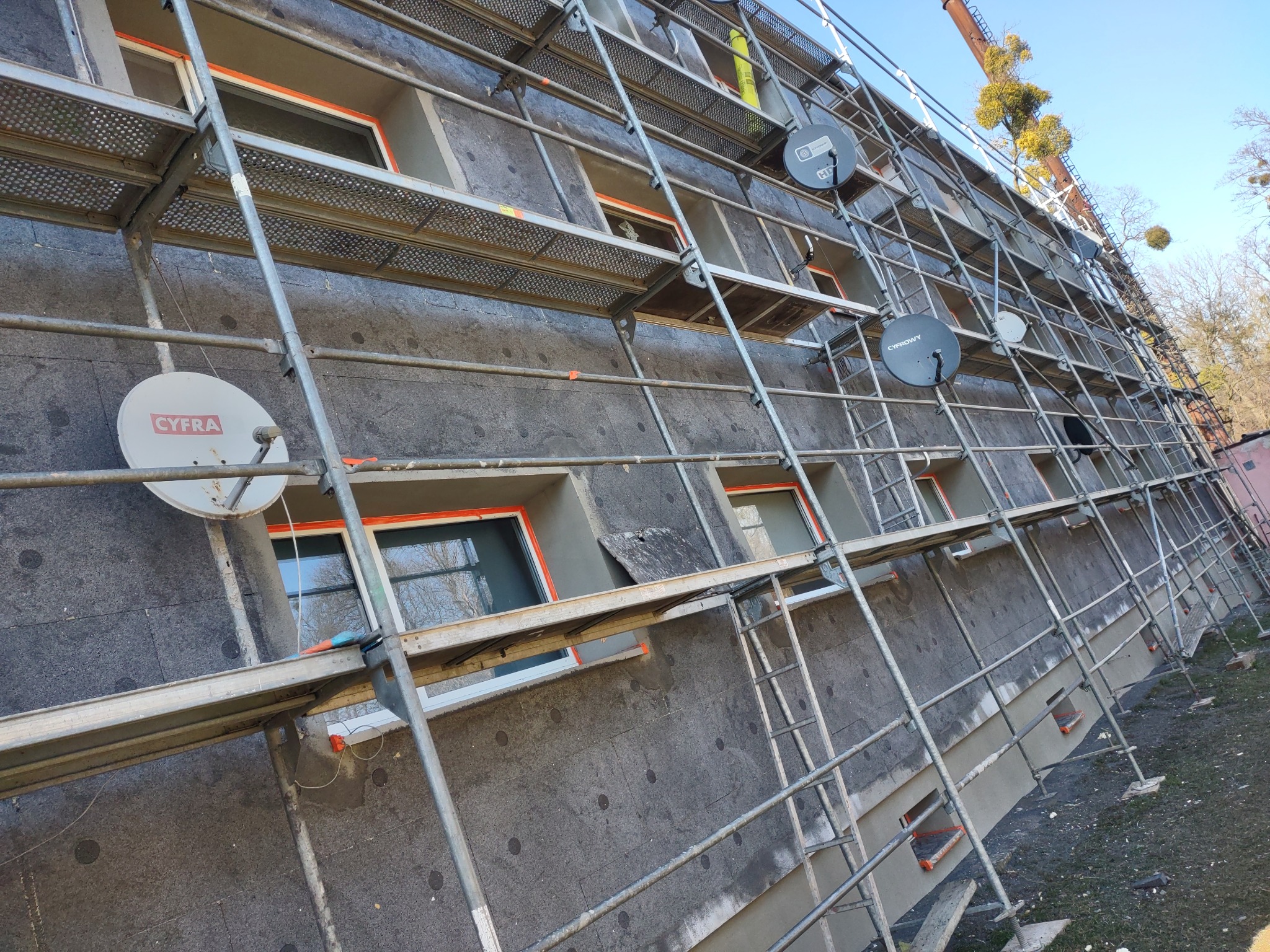 Budynek w trakcie ocieplania, widoczne rusztowania, styropian i okna z pomarańczowymi ramkami. Na elewacji zamontowane anteny satelitarne.
