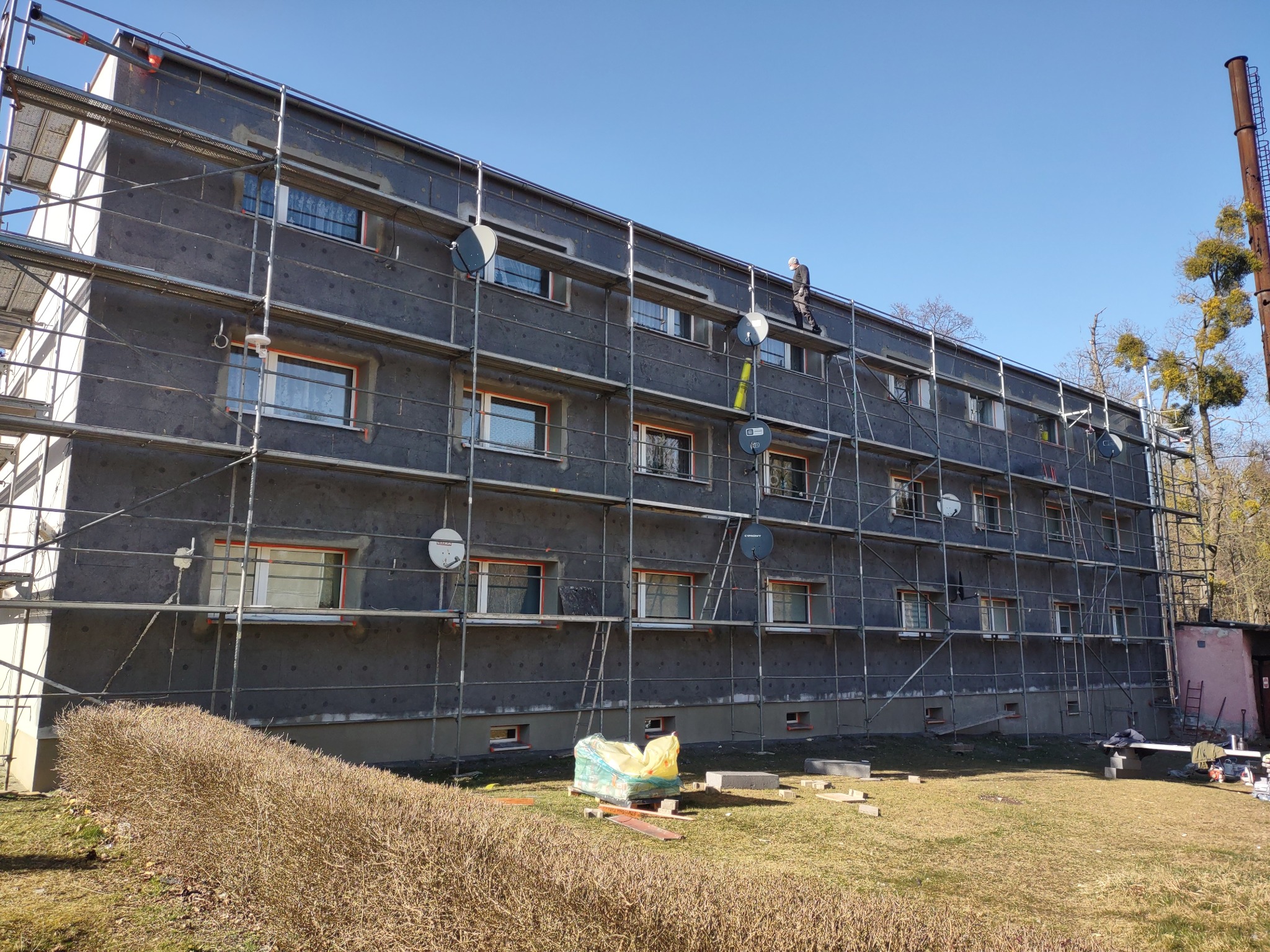 Budynek wielorodzinny w trakcie ocieplania w Strzelcach Opolskich, z widocznymi rusztowaniami i pracownikiem na wysokości. Elewacja pokryta szarą warstwą izolacyjną.