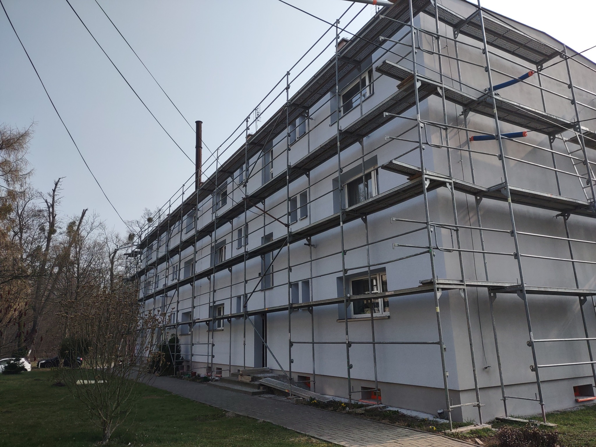 Wielopiętrowy budynek w rusztowaniach po malowaniu elewacji, widoczny komin i fragment drzew. Jasne, dzienne światło.