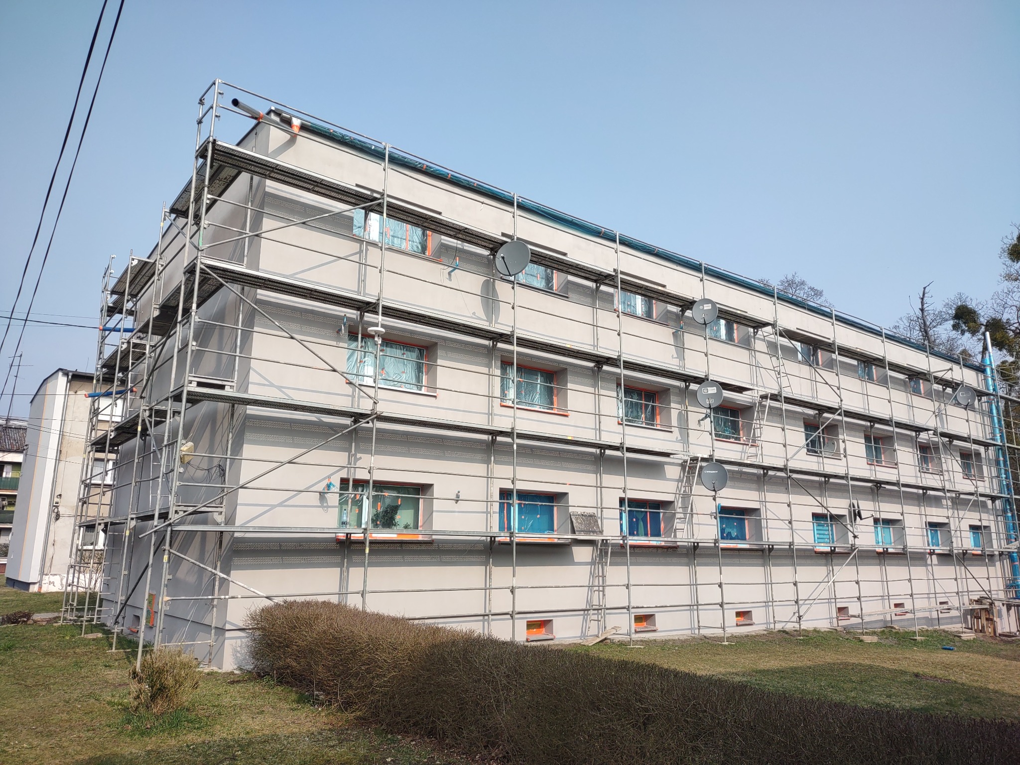 Wielopiętrowy budynek w trakcie prac elewacyjnych, osłonięte okna folią, otoczony rusztowaniami. Widoczne satelity na elewacji. Jasne, dzienne światło.