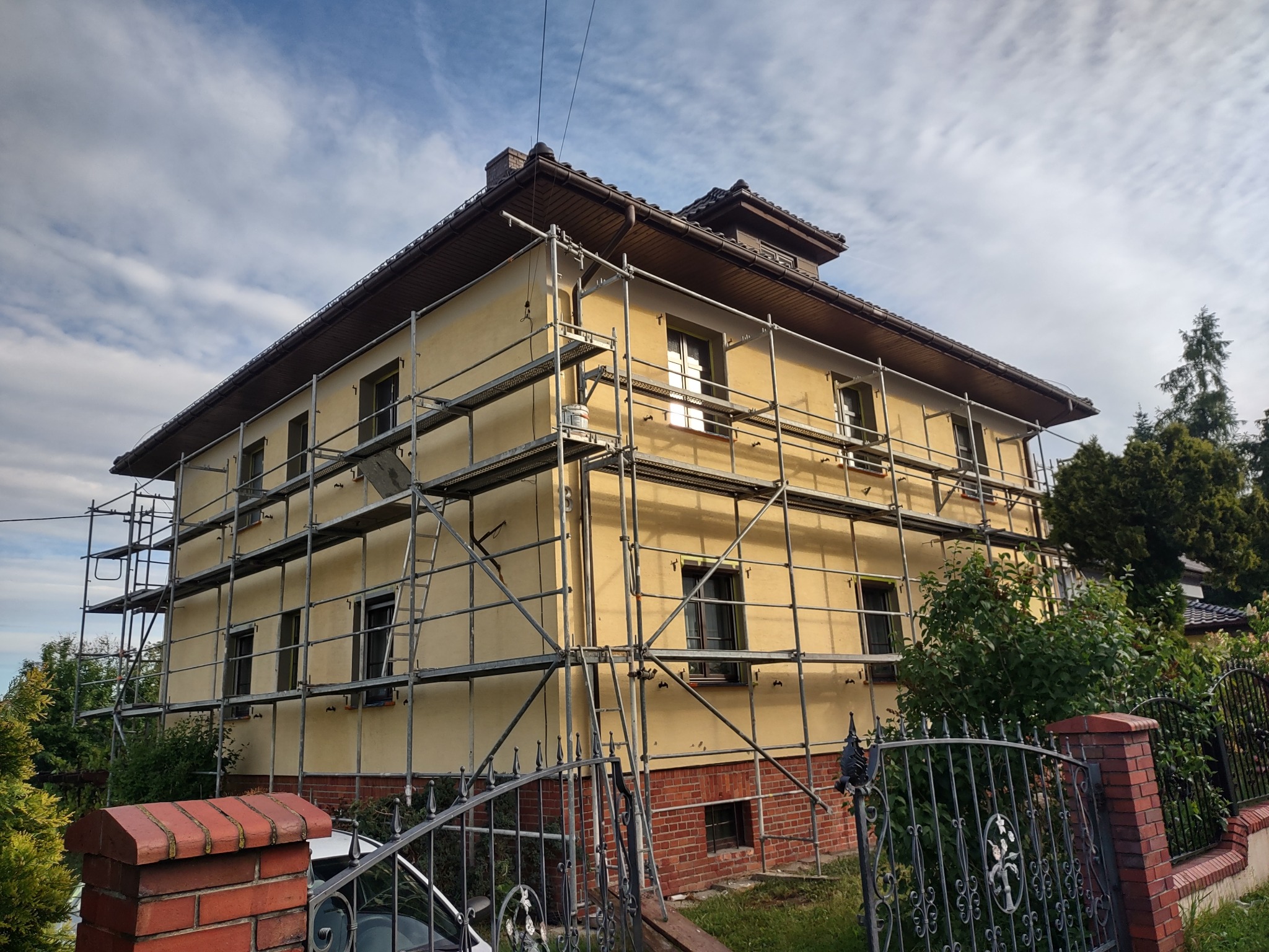 Żółta elewacja domu w rusztowaniach, widok zza ozdobnego, metalowego ogrodzenia. Częściowo widoczny ceglany cokół budynku. Błękitne niebo w tle.
