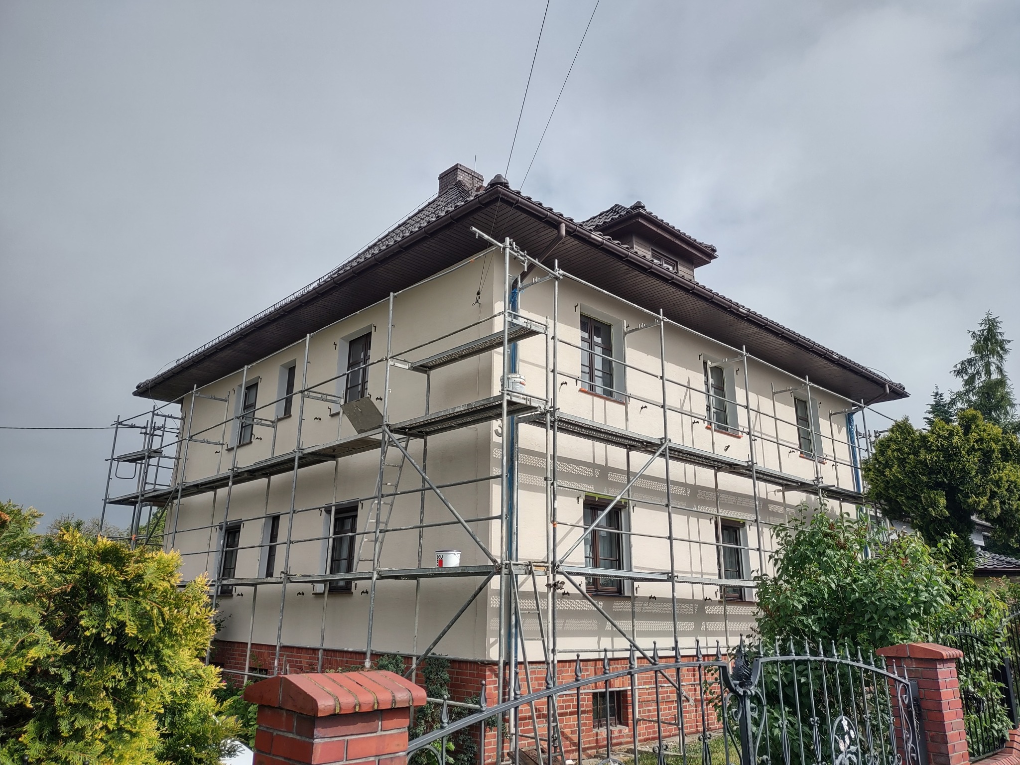 Dwupiętrowy dom w rusztowaniach podczas malowania elewacji na kremowo, z widocznym wiadrem farby. Częściowo pomalowana ściana i pracownik na rusztowaniu.