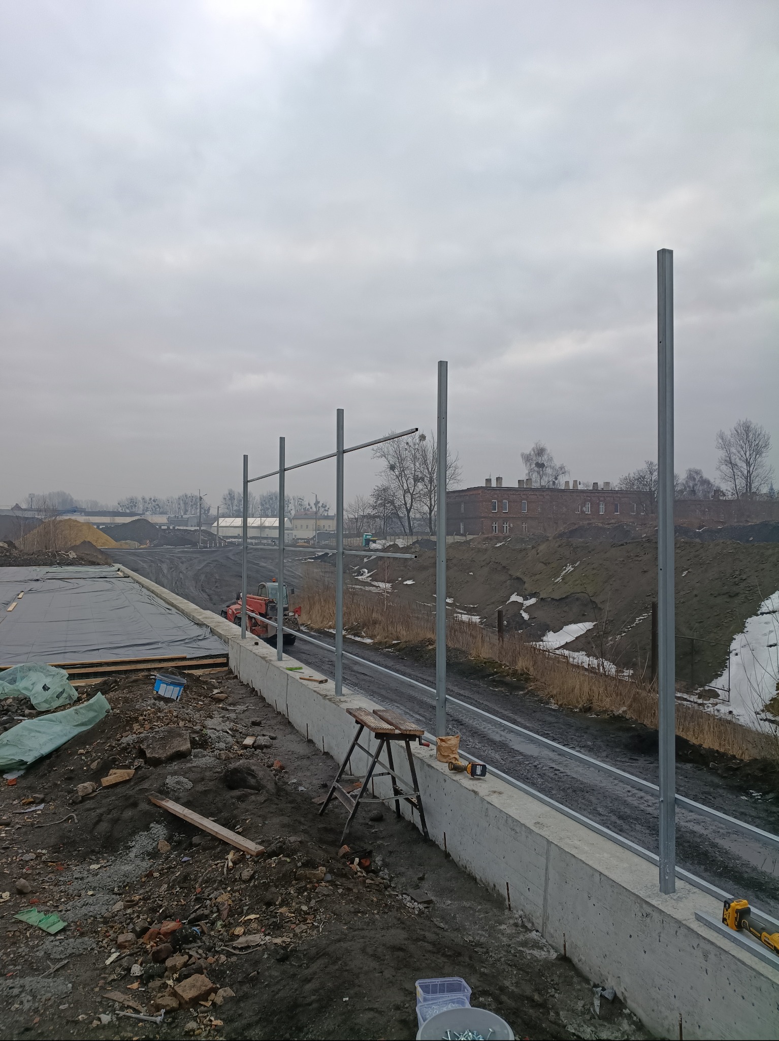 Wznoszenie betonowego fundamentu z wystającymi prętami zbrojeniowymi. W tle widoczny teren budowy, budynek i pochmurne niebo.