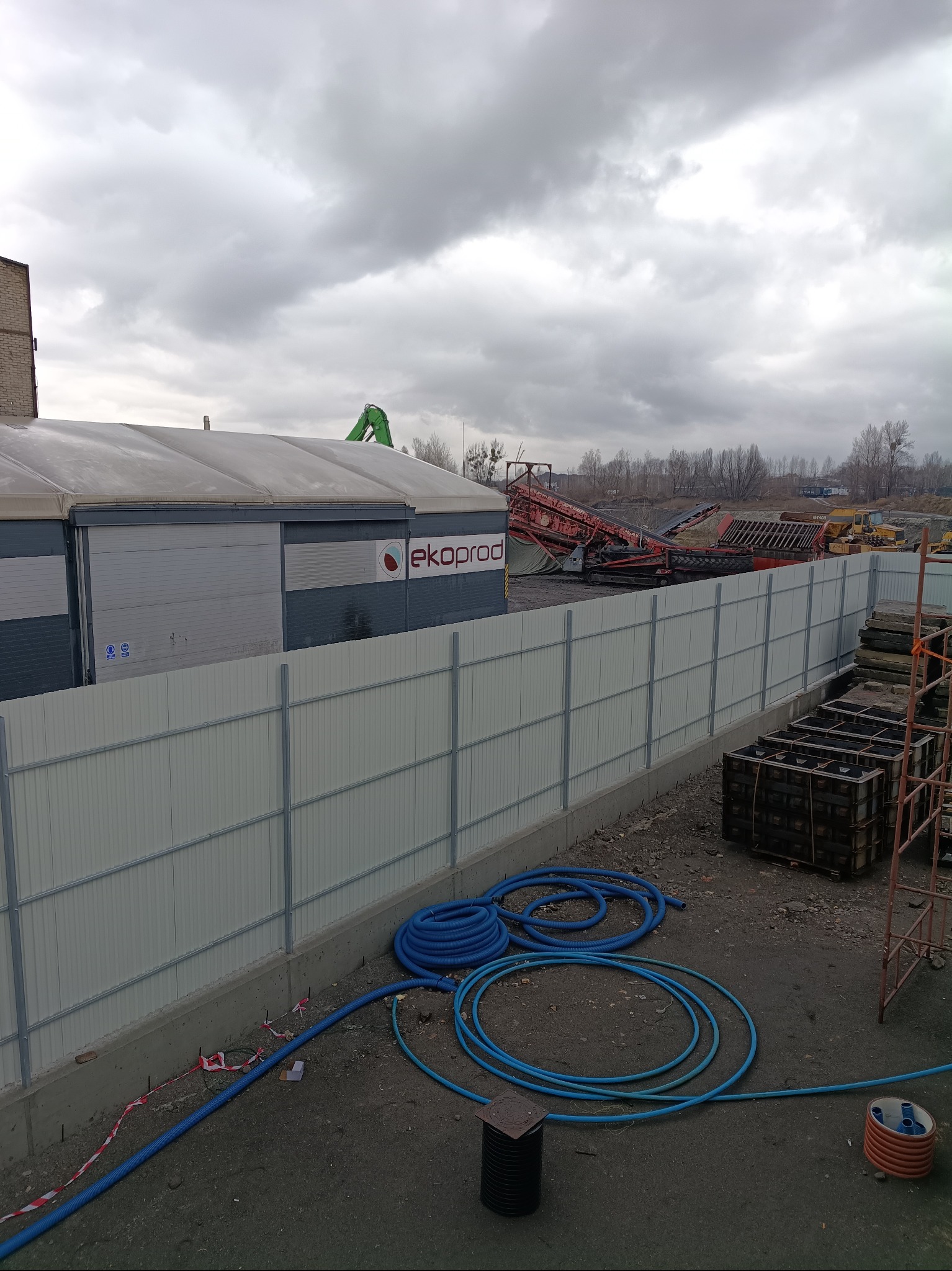 Teren budowy z nowym ogrodzeniem z blachy trapezowej, widoczne niebieskie rury, szalunki i maszyny budowlane w tle pod pochmurnym niebem.