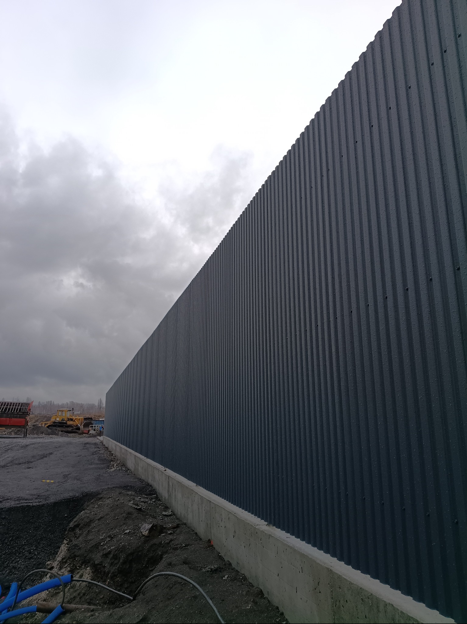Długa, szara ściana z blachy falistej na betonowej podmurówce, widoczna z boku, z ziemią i sprzętem budowlanym w tle pod pochmurnym niebem.