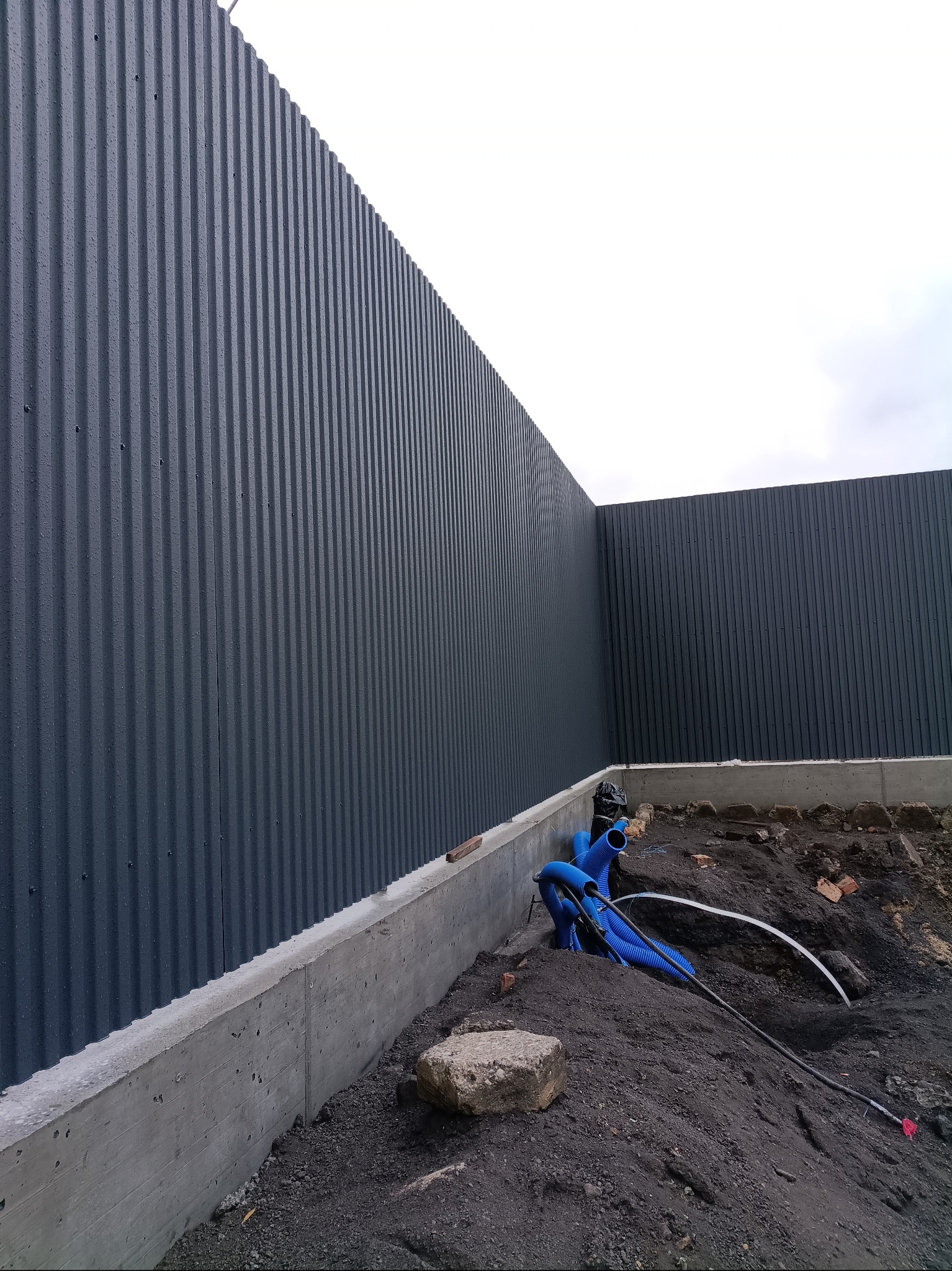 Szara, blaszana elewacja budynku z widocznymi fundamentami i instalacją w ziemi. Ujęcie z dołu, podkreślające pionowe linie blachy i surowość betonu.