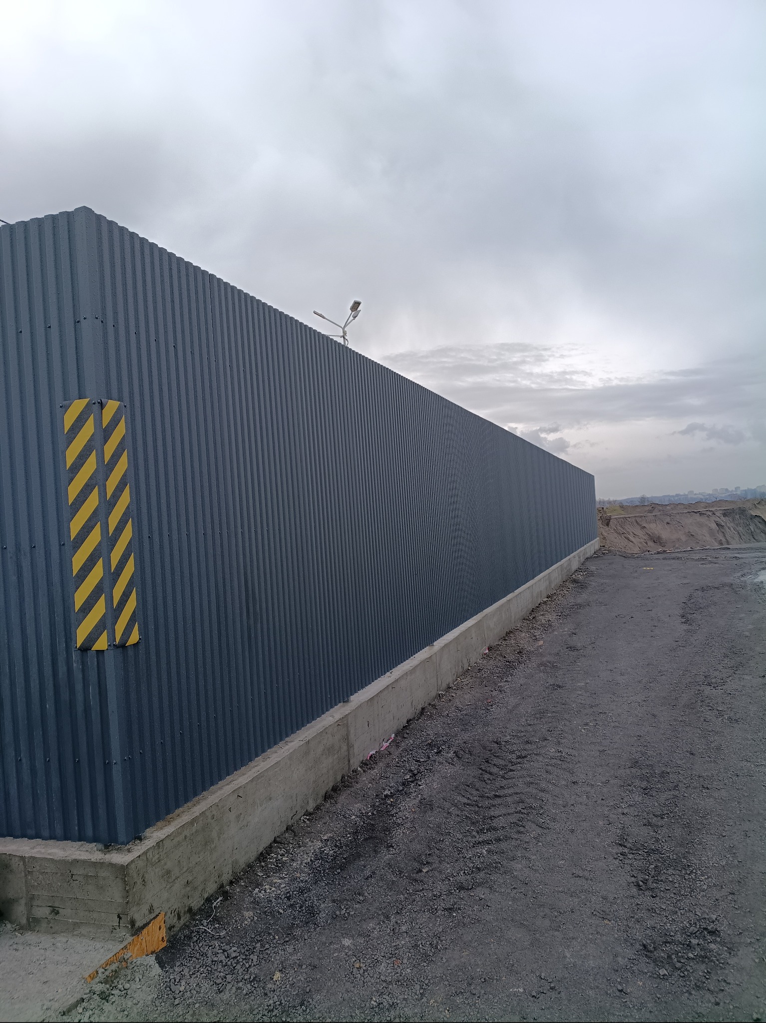 Długi, szary płot z blachy falistej na betonowej podmurówce, z żółto-czarnymi pasami ostrzegawczymi, oświetlony lampą, na tle pochmurnego nieba i hałdy ziemi.