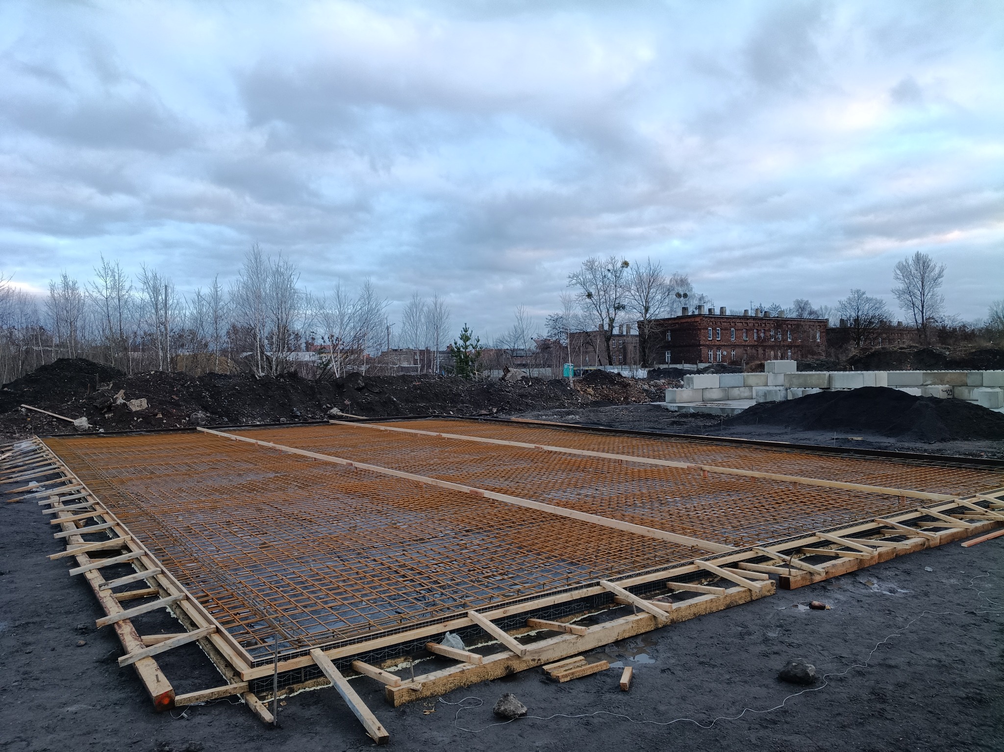 Siatka zbrojeniowa ułożona w drewnianym szalunku na przygotowanym terenie budowy, w tle budynek z czerwonej cegły i pochmurne niebo.
