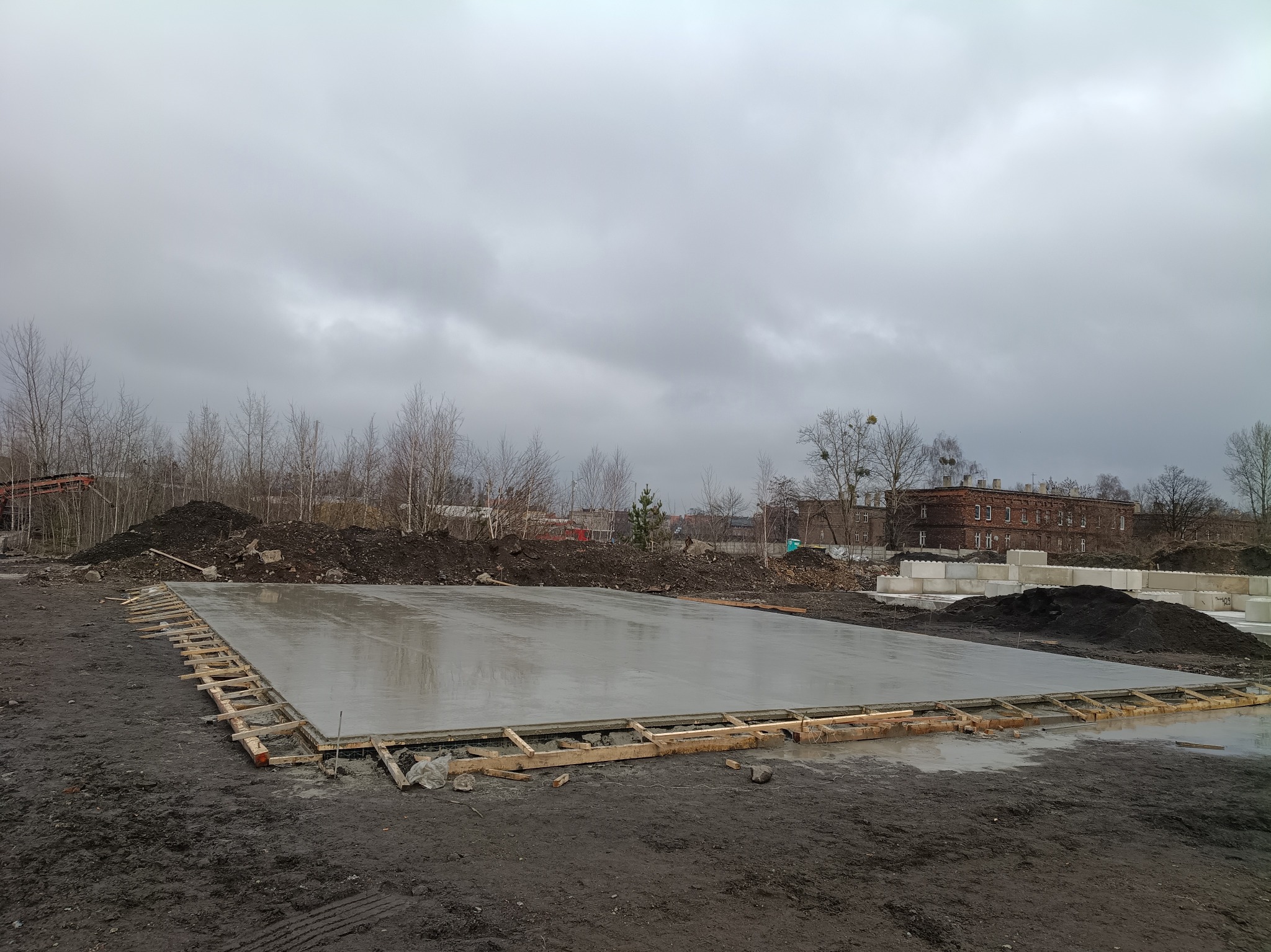 Świeżo wylana, gładka płyta fundamentowa z drewnianym szalunkiem na błotnistym terenie. W tle hałda ziemi, budynek i pochmurne niebo.