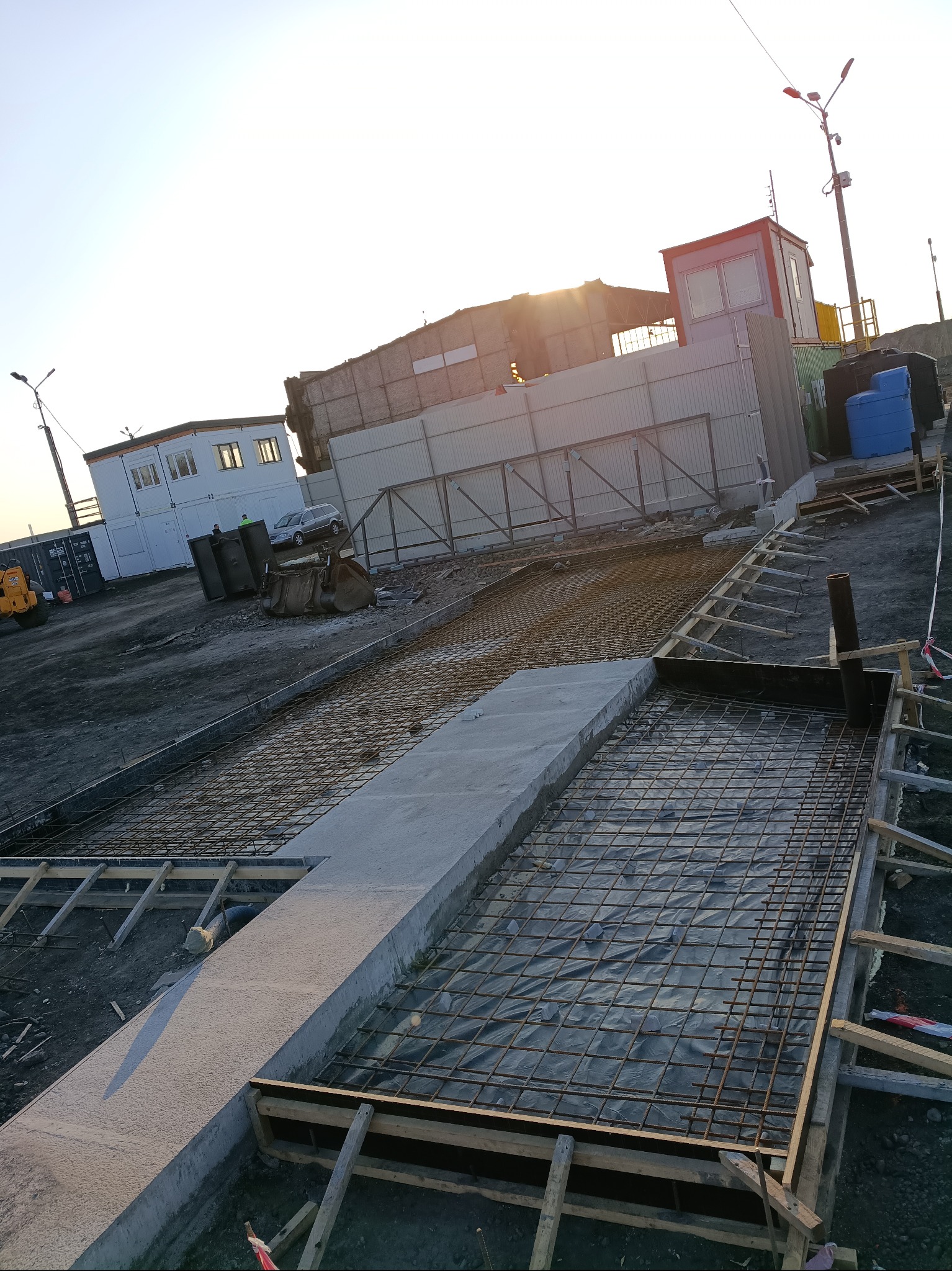 Zbrojenie fundamentów pod budynek, widoczne stalowe pręty i drewniane szalunki. W tle budynki i sprzęt budowlany w promieniach zachodzącego słońca.