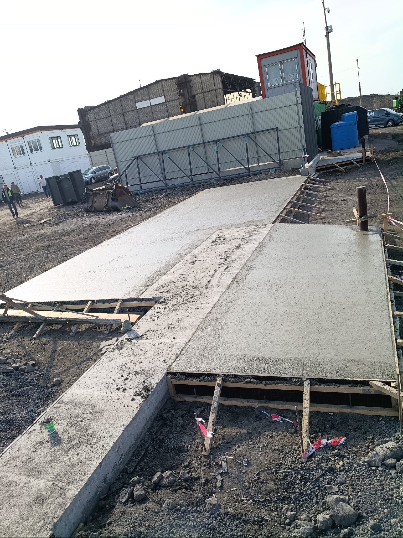 Wylany beton na przygotowanym szalunku. Widoczne drewniane elementy konstrukcji i świeży beton. W tle budynki i sprzęt budowlany. Ujęcie z góry.