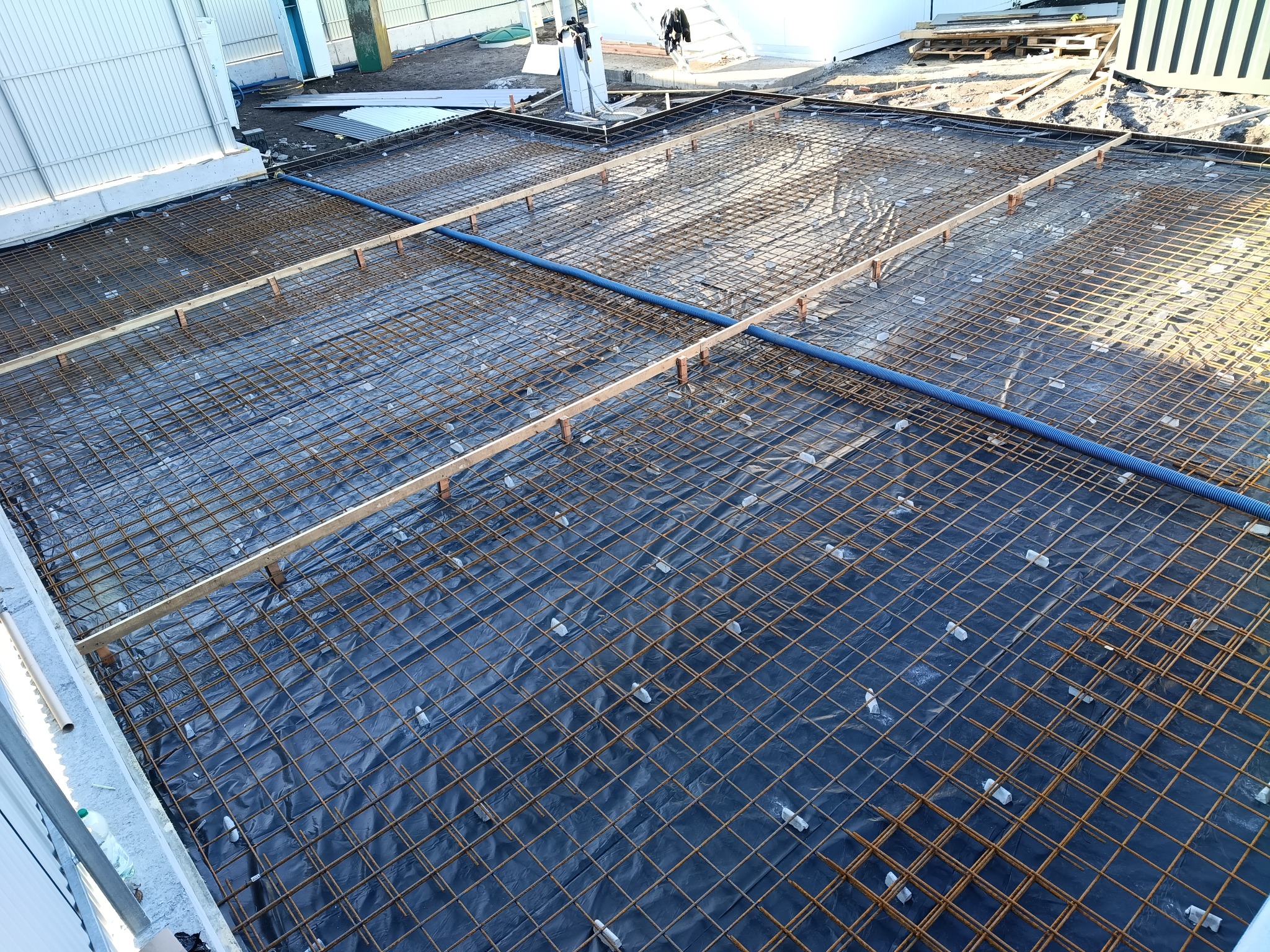 Siatka zbrojeniowa na czarnej folii, podparta drewnianymi belkami i plastikowymi dystansami, przygotowana do zalania betonem. Widok z góry na plac budowy.