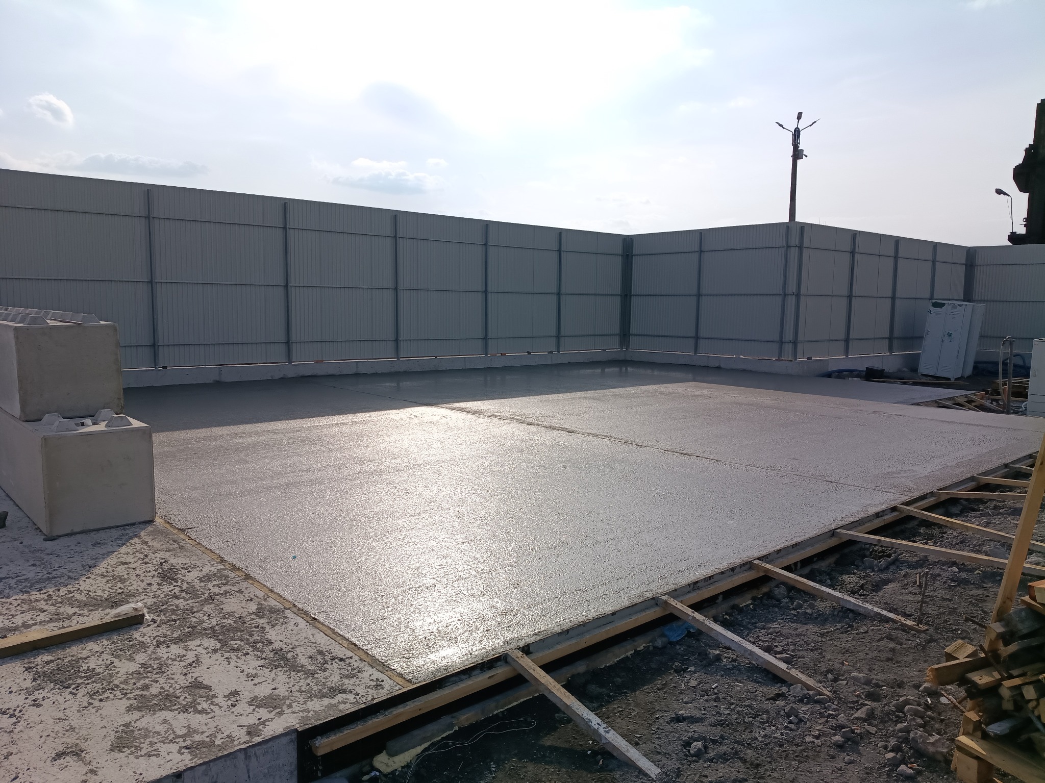 Świeżo wylany, mokry beton na przygotowanym fundamencie z drewnianym szalunkiem, otoczony szarą, metalową konstrukcją. Słoneczny dzień.