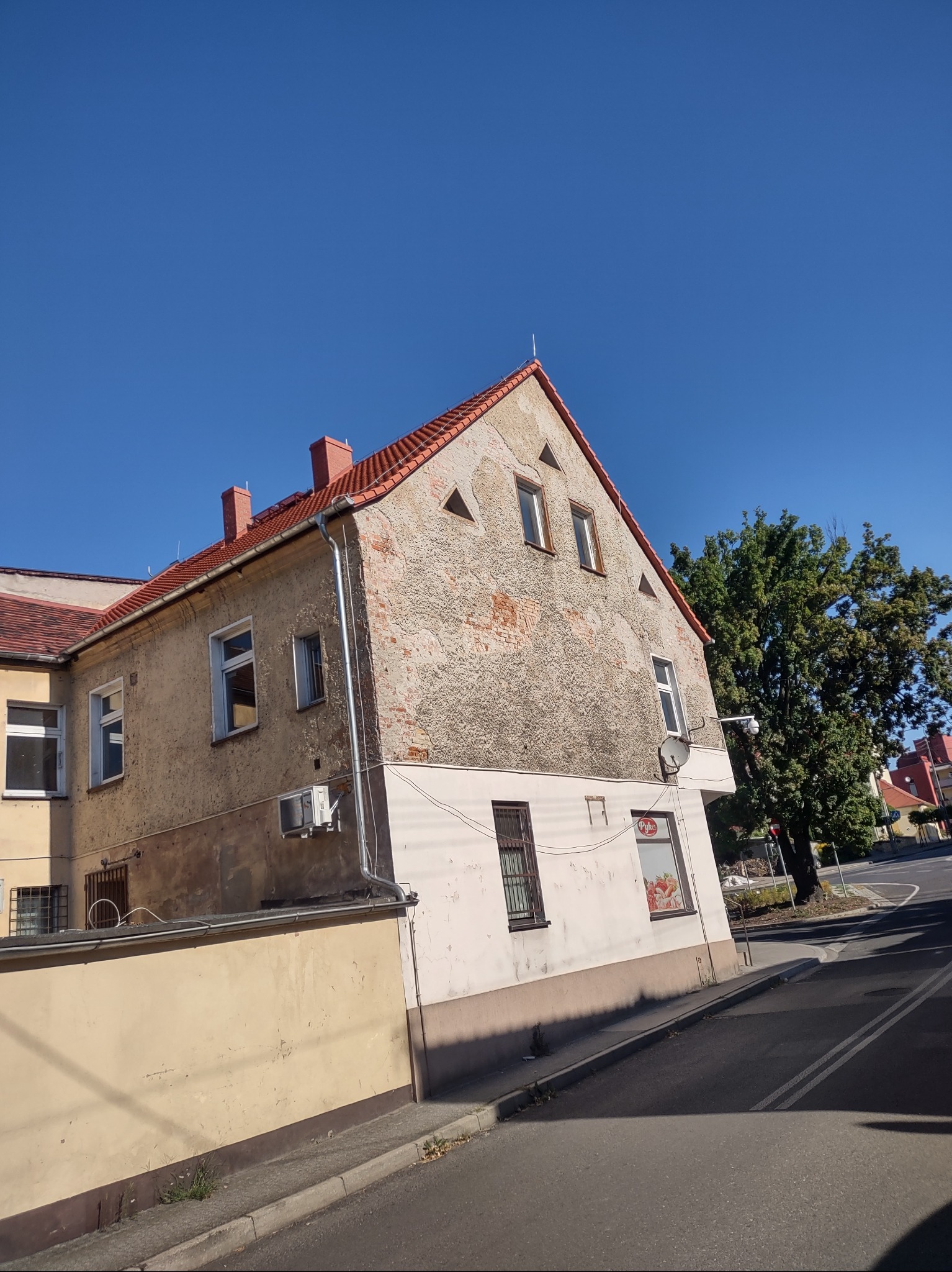 Dwukondygnacyjny budynek z nierówną elewacją, częściowo odnowiony, z czerwonym dachem i kominami, widziany z poziomu ulicy w słoneczny dzień.