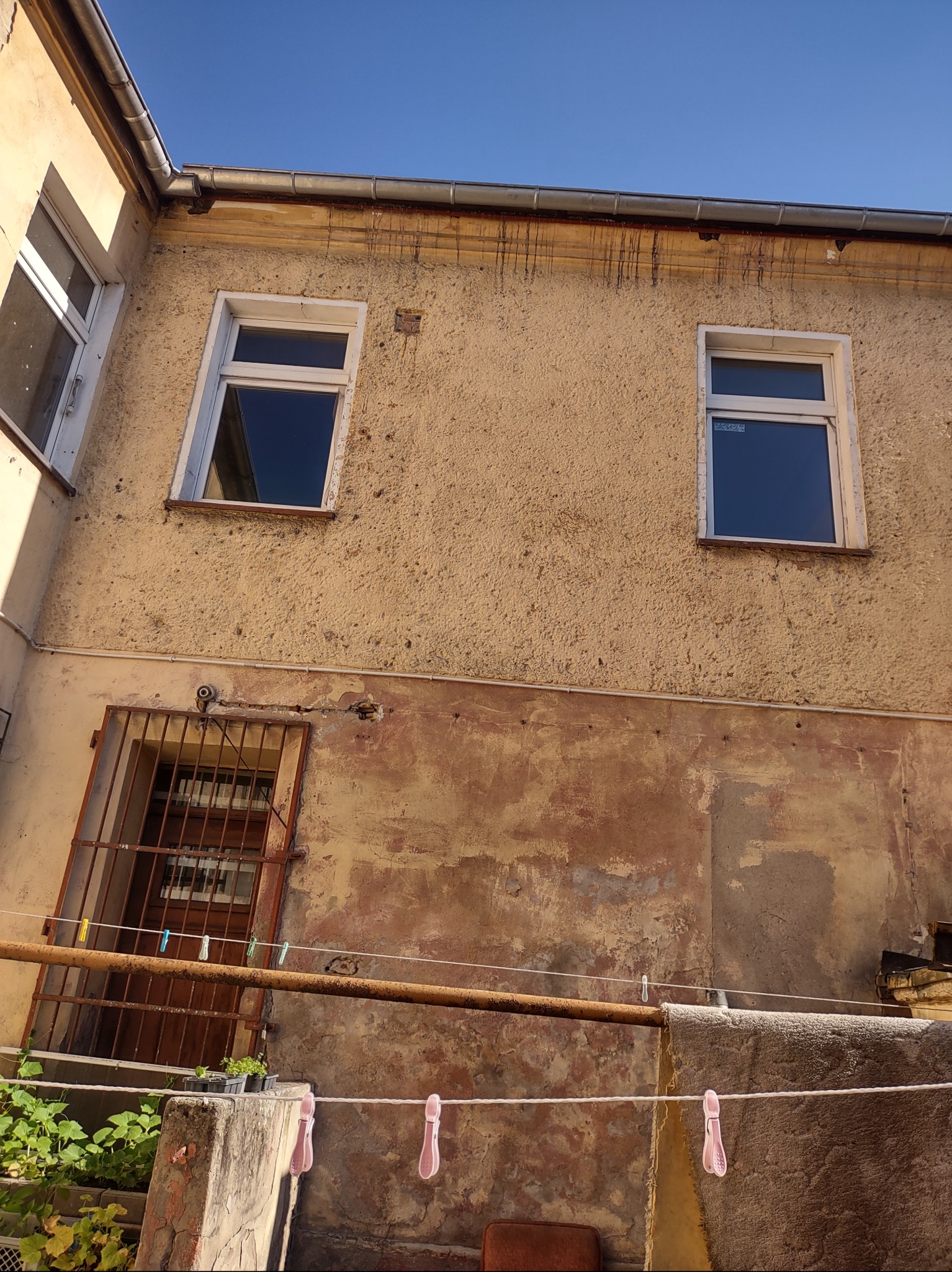 Zniszczona elewacja budynku z dwoma oknami i kratą. Widoczne ślady zacieków i odpadającego tynku. Na pierwszym planie suszarka z praniem i rośliny w doniczkach.