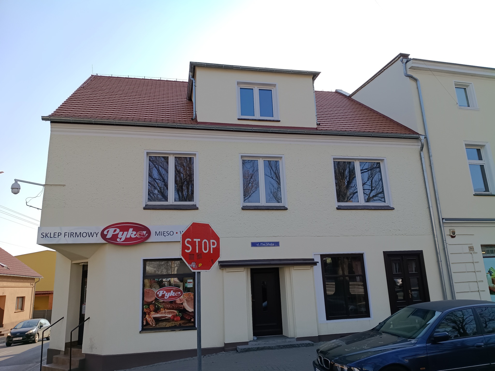 Odnowiona elewacja budynku sklepu w Strzelcach Opolskich. Jasna fasada z czerwonym dachem, okna z odbiciami drzew. Widoczny szyld sklepu i znak STOP.