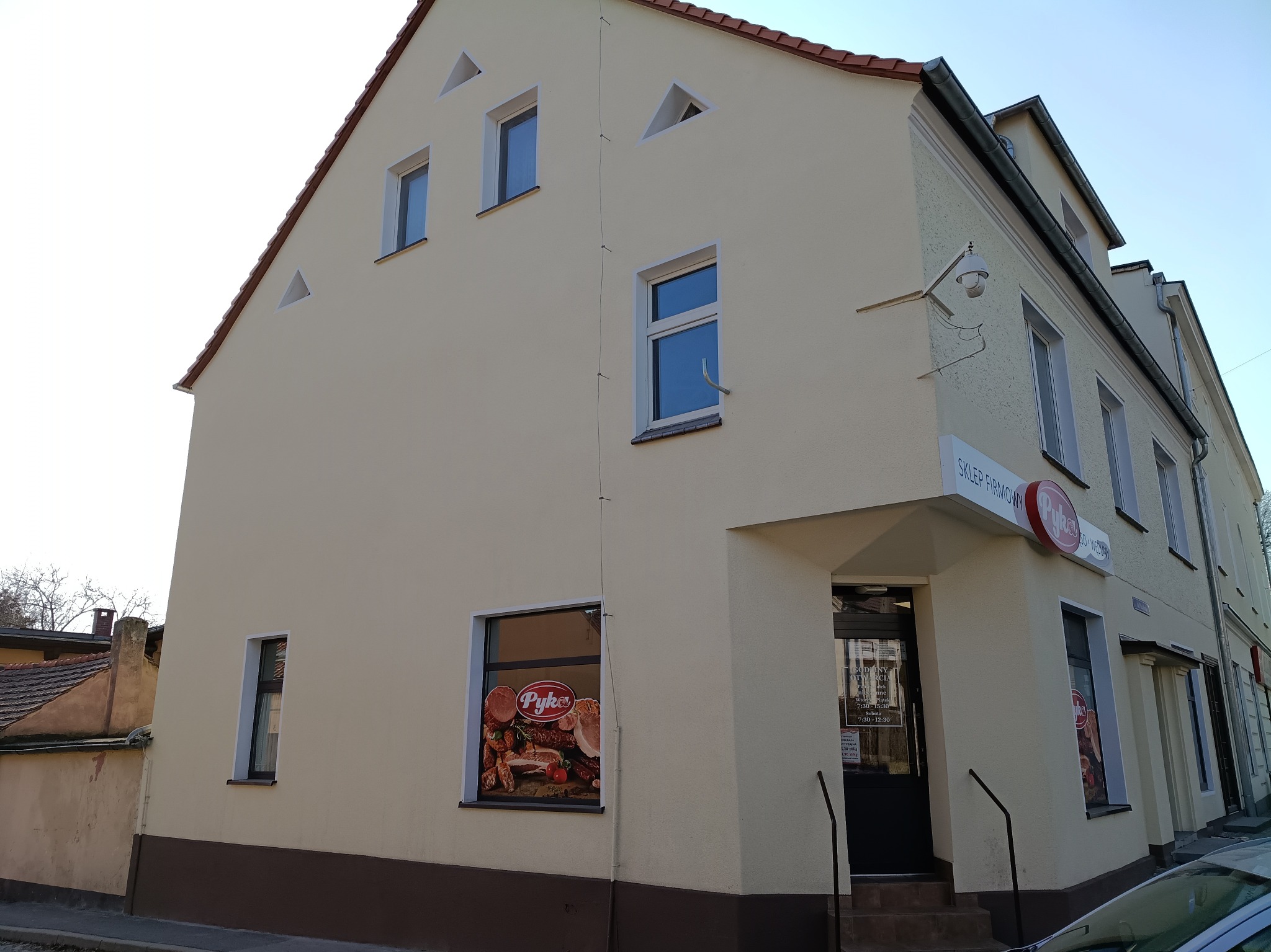 Jasna elewacja budynku sklepu z brązowym pasem u dołu. Widoczne okna, drzwi wejściowe i szyld sklepu. Ujęcie z dołu, podkreślające wysokość budynku.