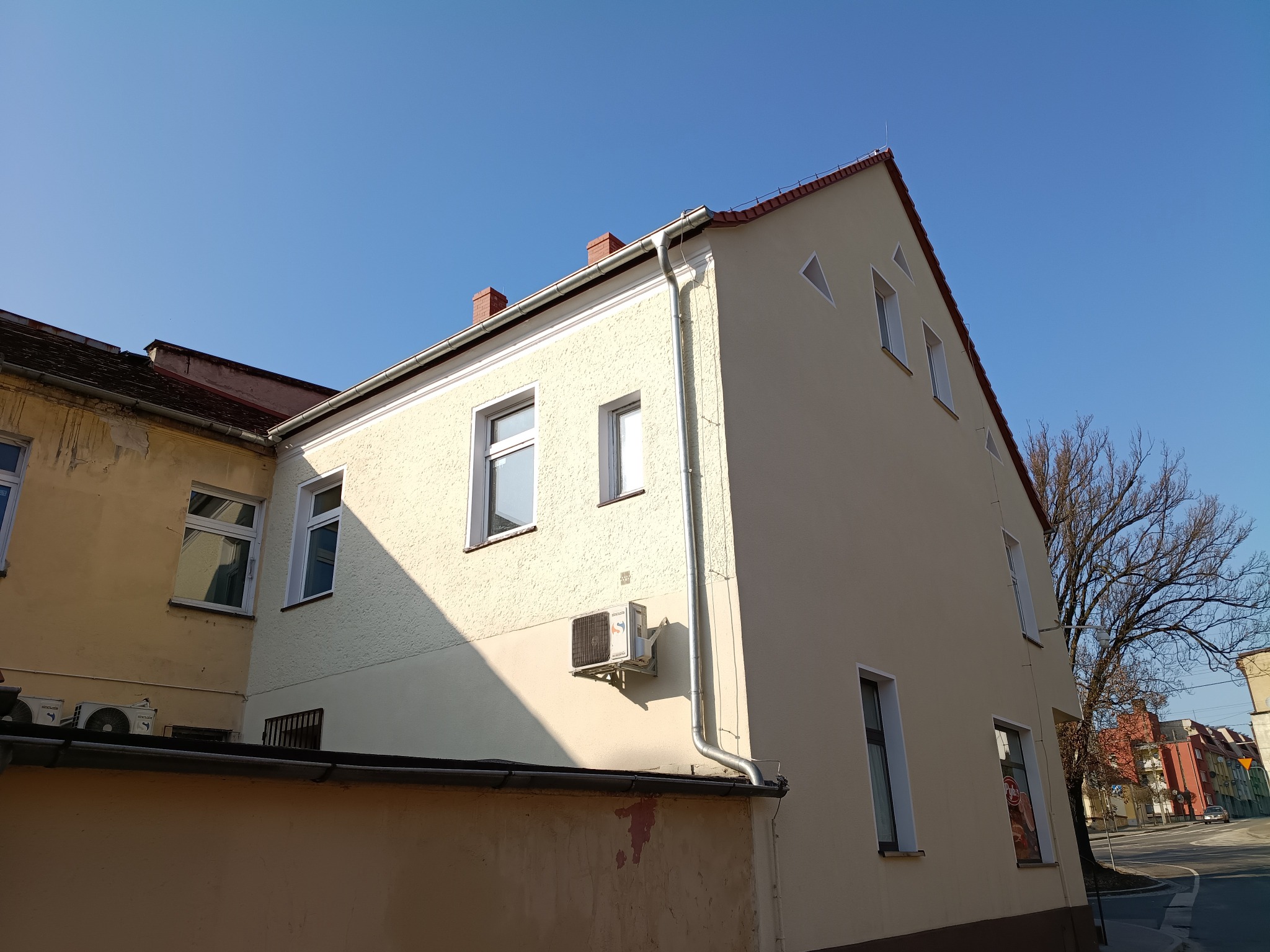 Odnowiona elewacja budynku z klimatyzatorem, widoczna z perspektywy ulicy w słoneczny dzień. Jasna, gładka fasada kontrastuje z sąsiednim budynkiem o starszej elewacji.