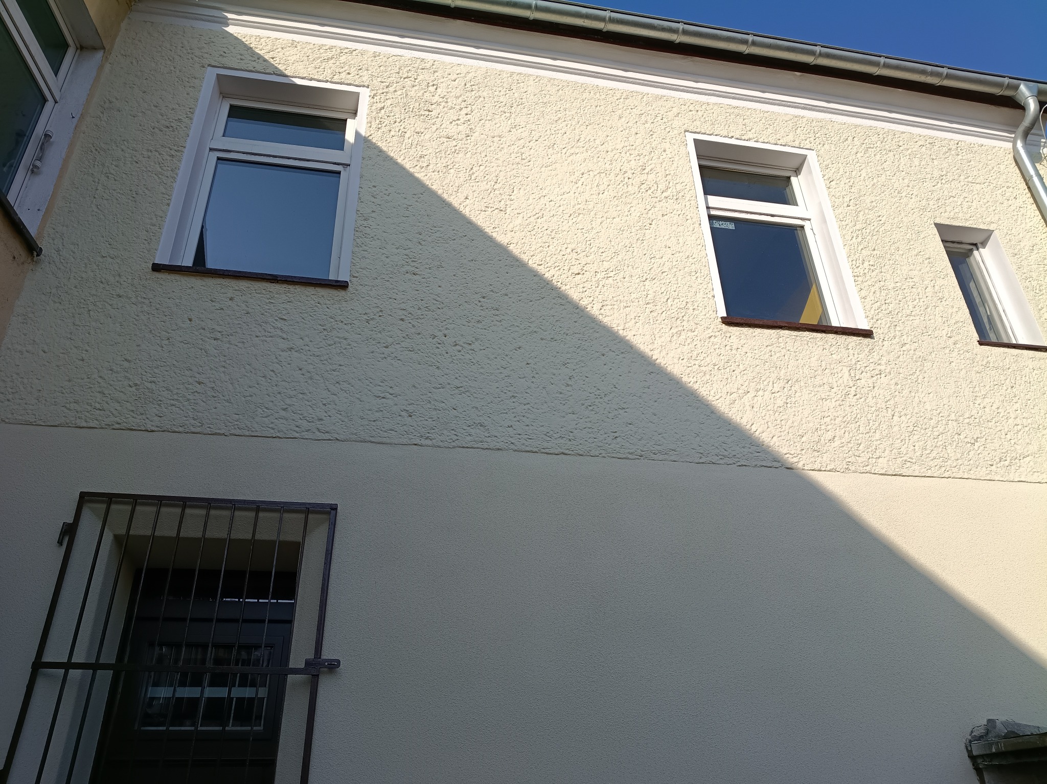 Jasna elewacja budynku z trzema oknami, z czego jedno z kratą. Na elewacji widoczny cień, a w tle błękitne niebo.