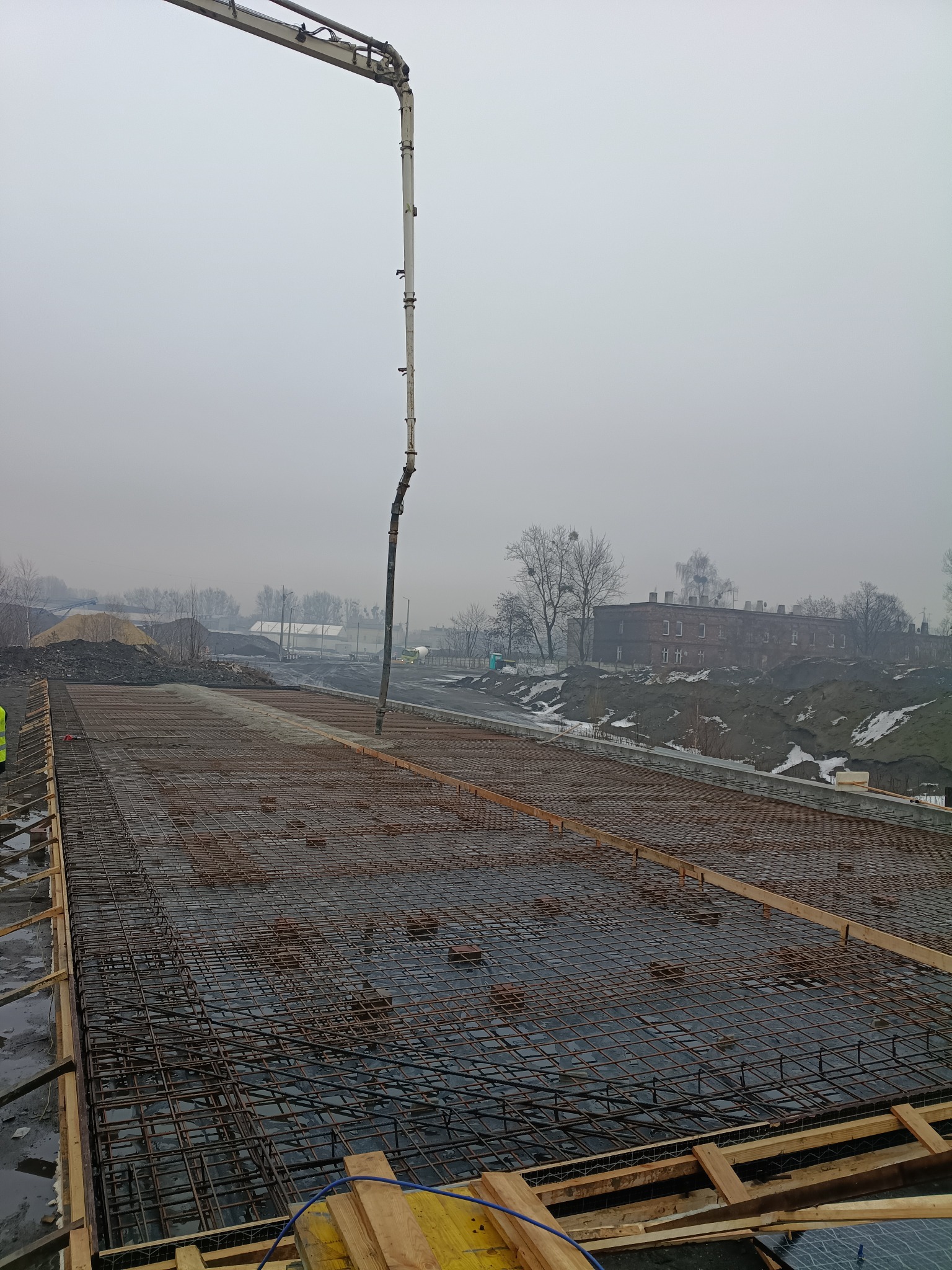 Zbrojenie fundamentów z użyciem pompy do betonu na tle mglistego krajobrazu miejskiego. Widoczne drewniane szalunki i stalowe pręty zbrojeniowe.
