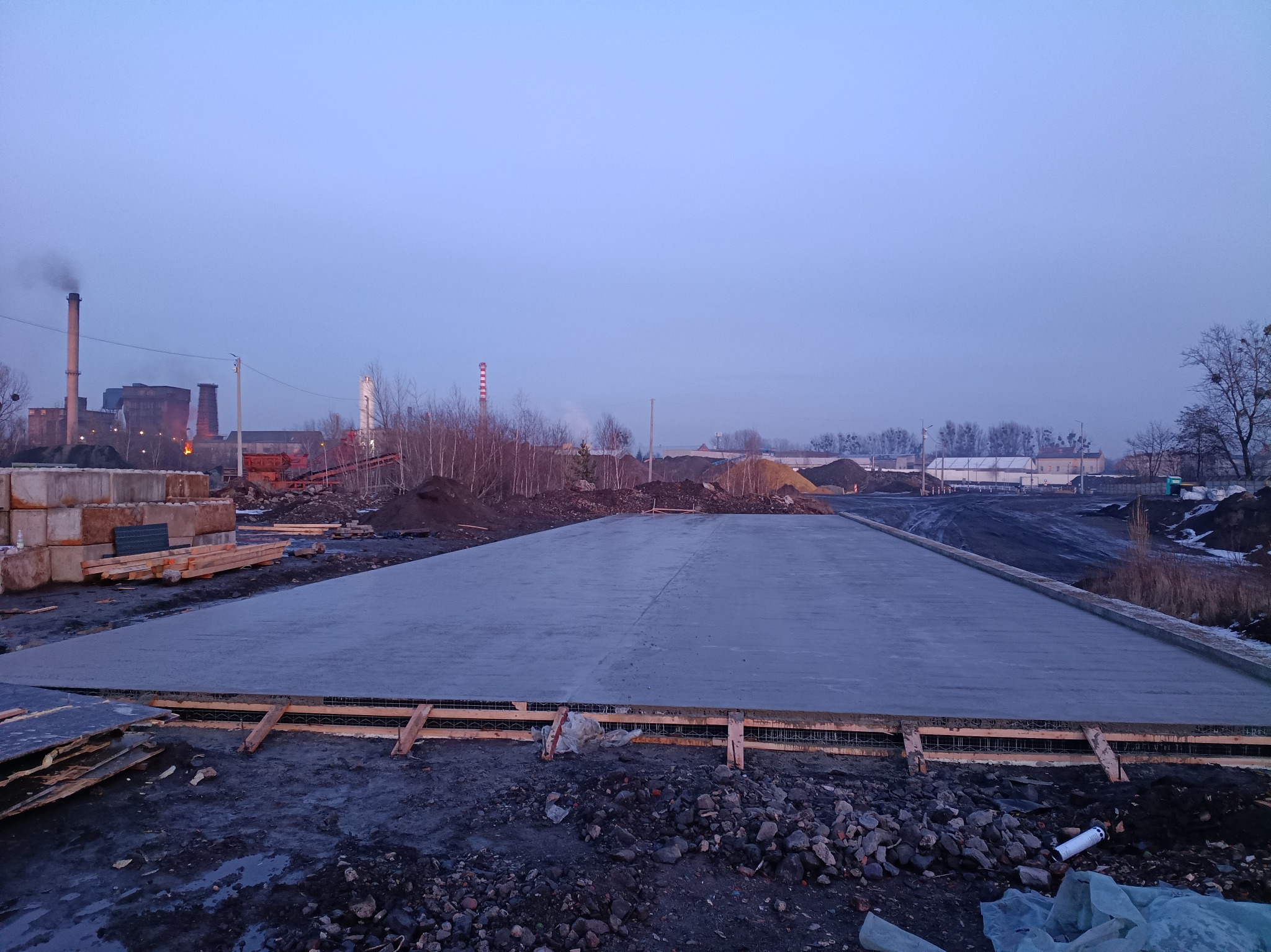 Wylana płyta fundamentowa z widocznym zbrojeniem i szalunkiem drewnianym. W tle kominy fabryczne i hałdy ziemi, zimowy krajobraz o zmierzchu.