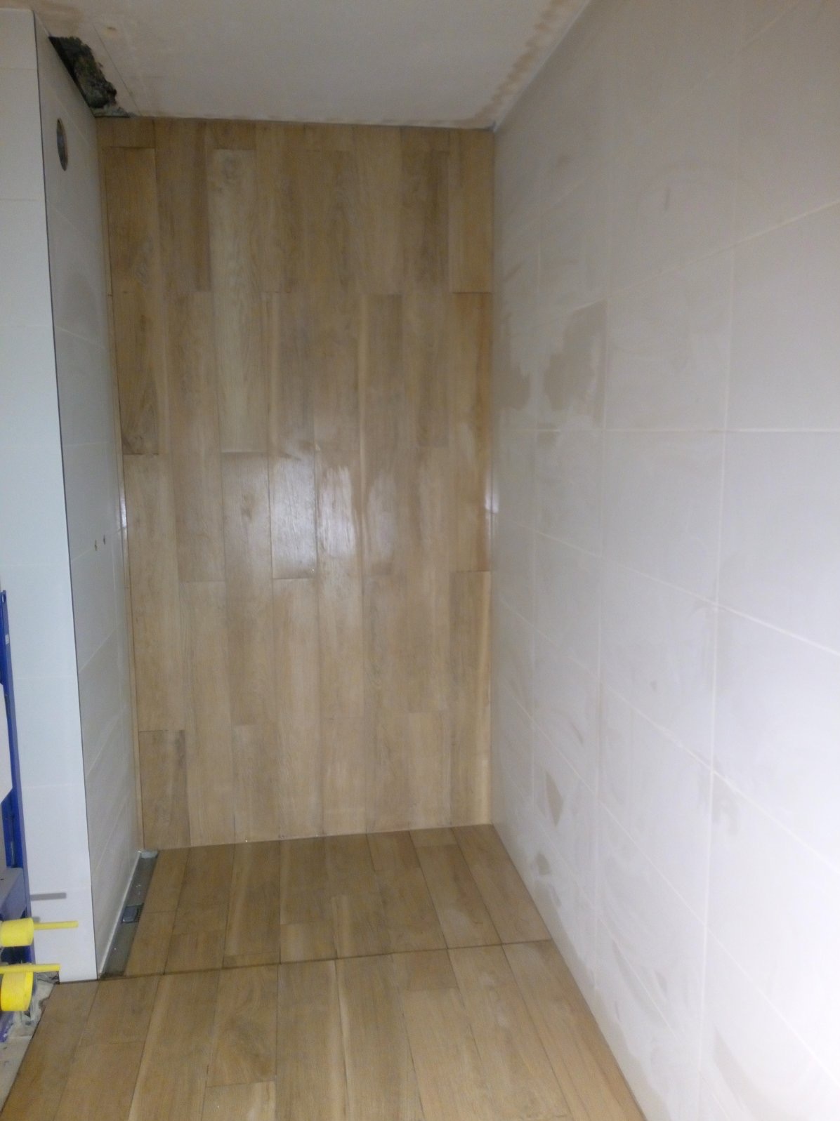 Niedokończona łazienka z drewnopodobnymi płytkami na podłodze i ścianie prysznica, białe płytki na pozostałych ścianach, widoczna instalacja podtynkowego WC.