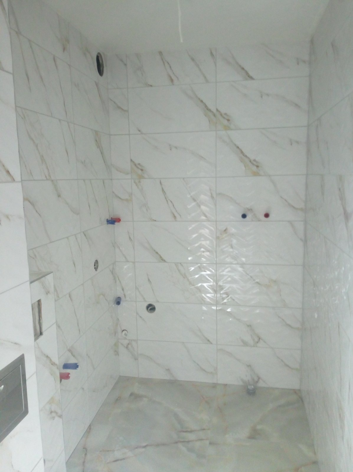 Wykończona łazienka z białymi płytkami imitującymi marmur, widoczne otwory na armaturę i odpływ pod prysznic bez brodzika.