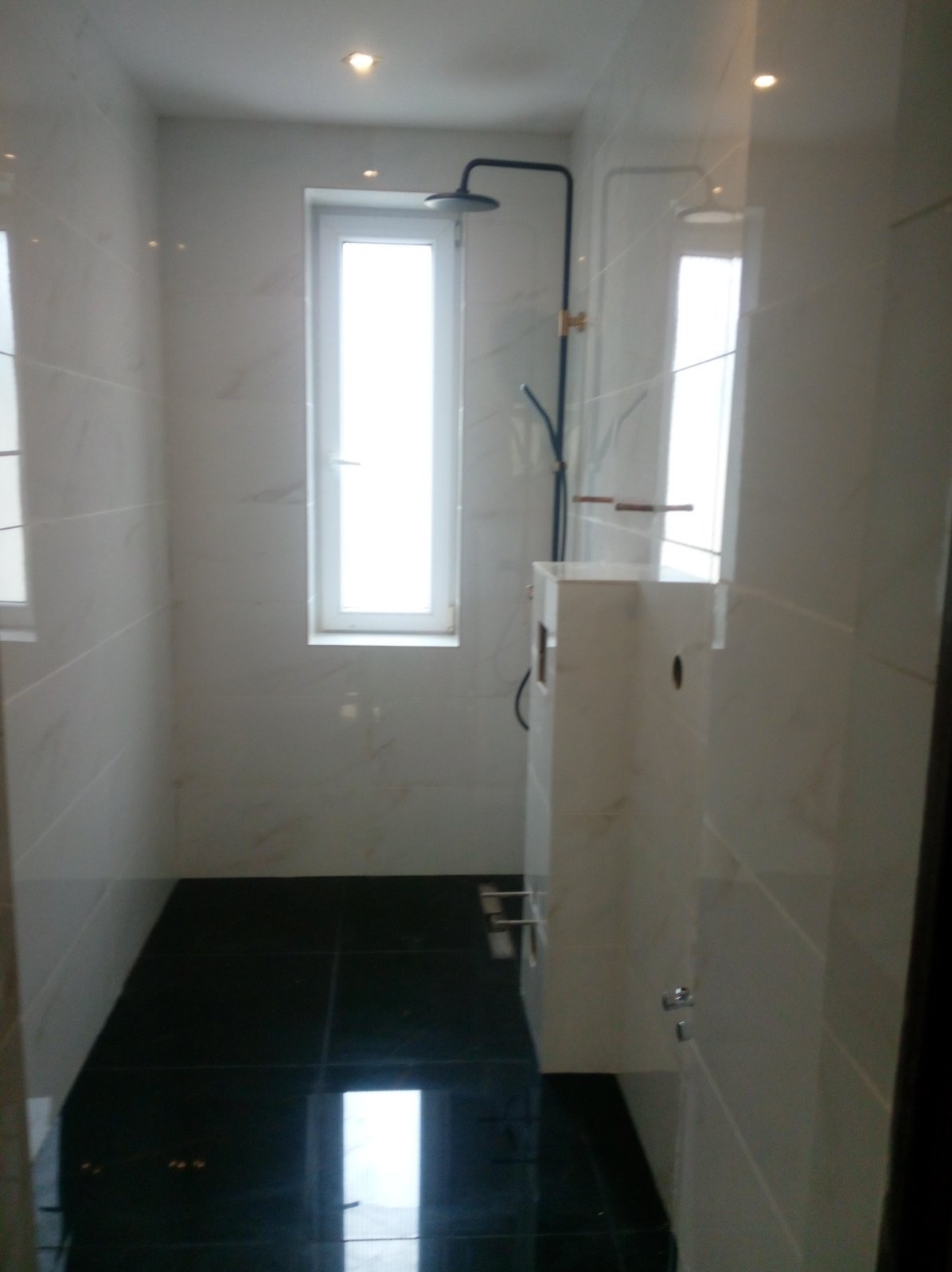 Wnętrze łazienki z czarną, błyszczącą podłogą, białymi marmurkowymi płytkami na ścianach, oknem w głębi i czarną deszczownicą.