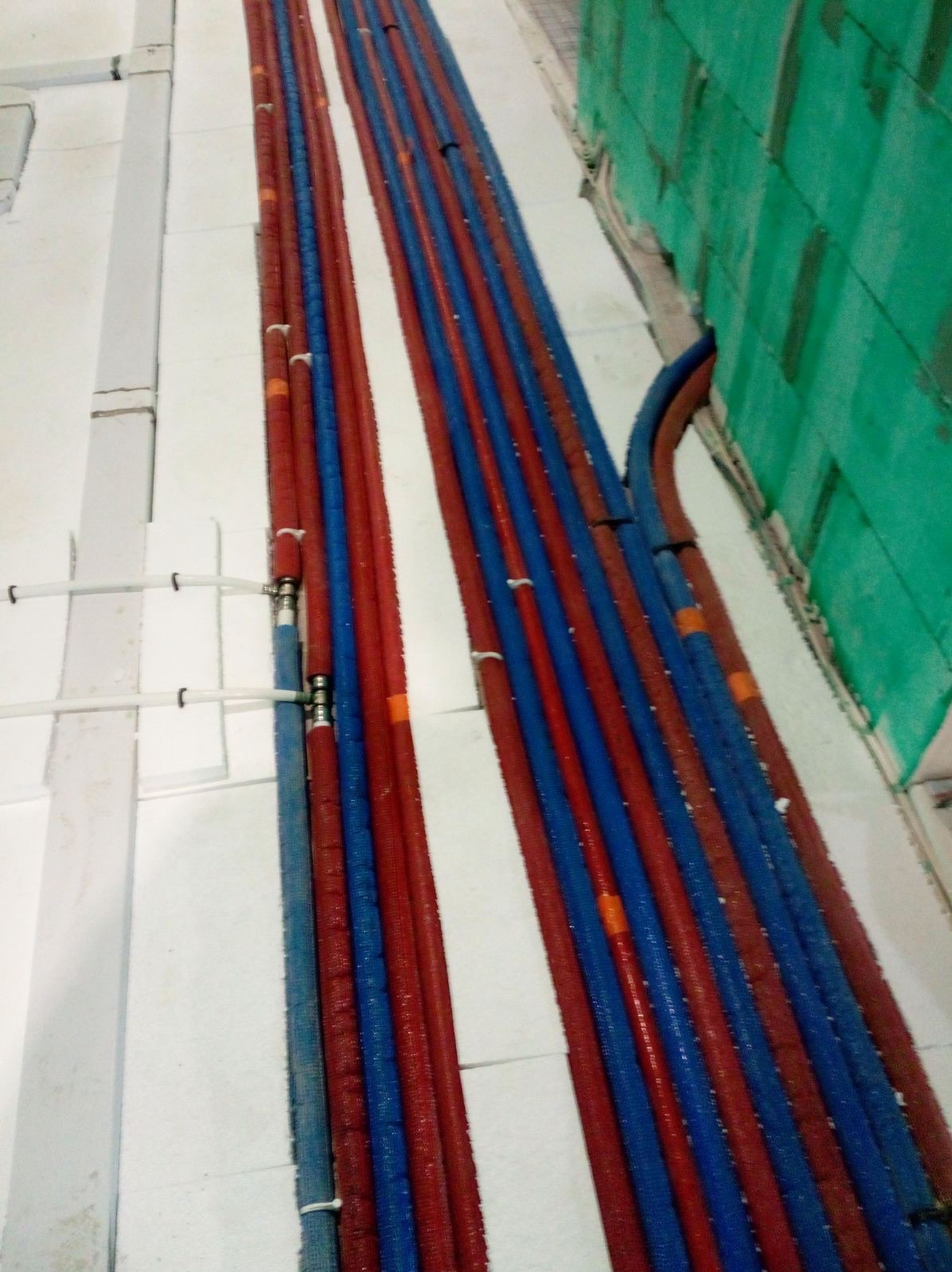 Równolegle ułożone, czerwono-niebieskie rury grzewcze z izolacją, przymocowane do białej ściany i podłogi w trakcie montażu instalacji.