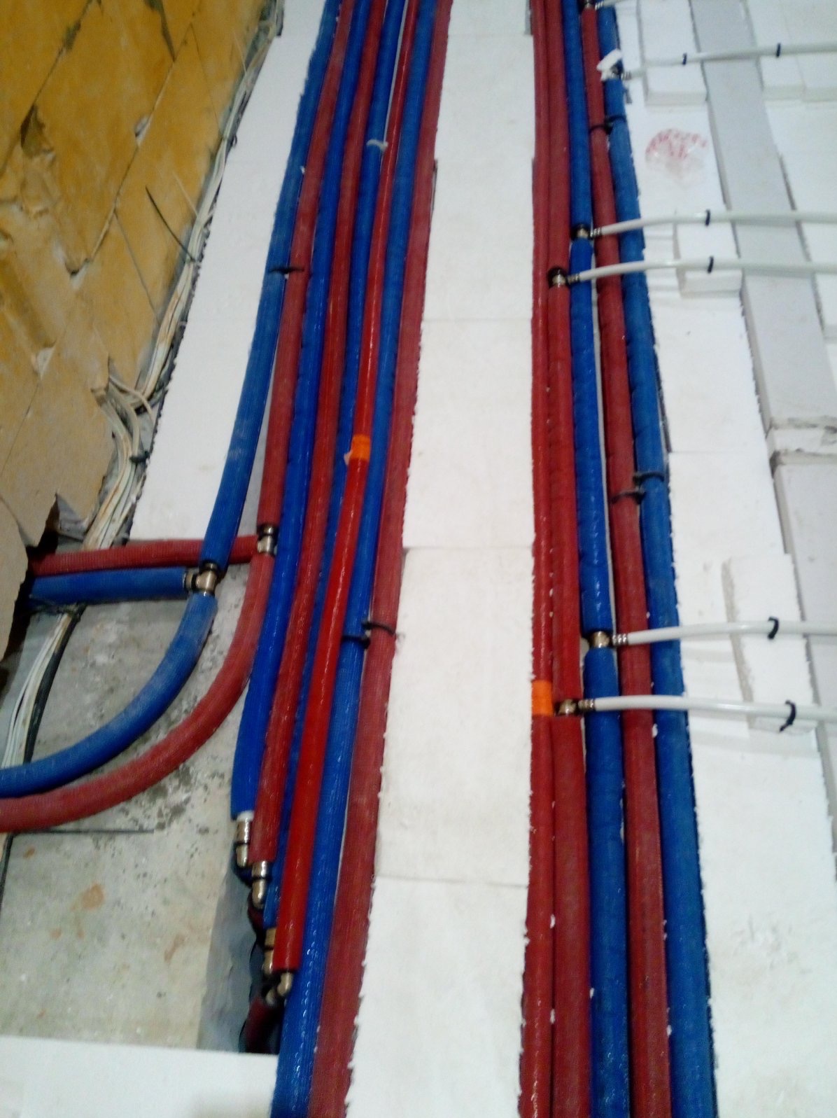 Instalacja podłogowa: czerwone i niebieskie rury grzewcze ułożone na białym styropianie, widoczne połączenia i fragment surowej ściany z przewodami elektrycznymi.