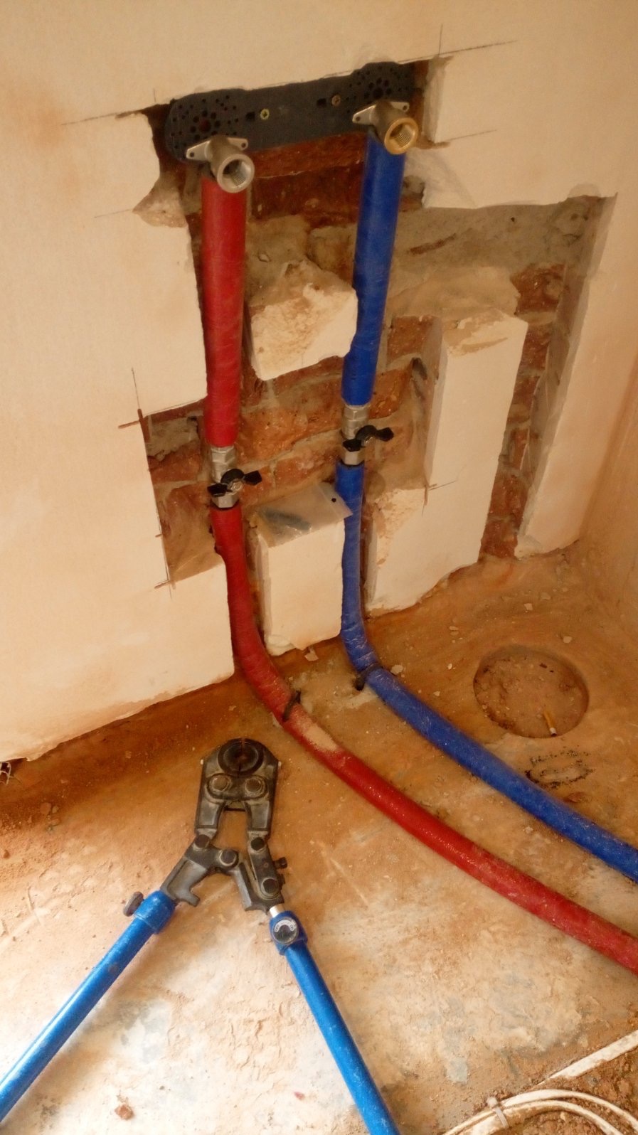 Montaż instalacji wodnej: czerwona i niebieska rura PEX z zaworami wychodzące ze ściany, obok leżą zaciskarki do rur, widoczne ślady po kuciu w ścianie.