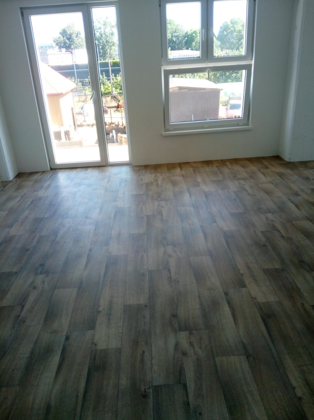 Nowo ułożone panele podłogowe w jasnym pomieszczeniu z dużymi oknami i drzwiami balkonowymi, widok na zewnątrz.