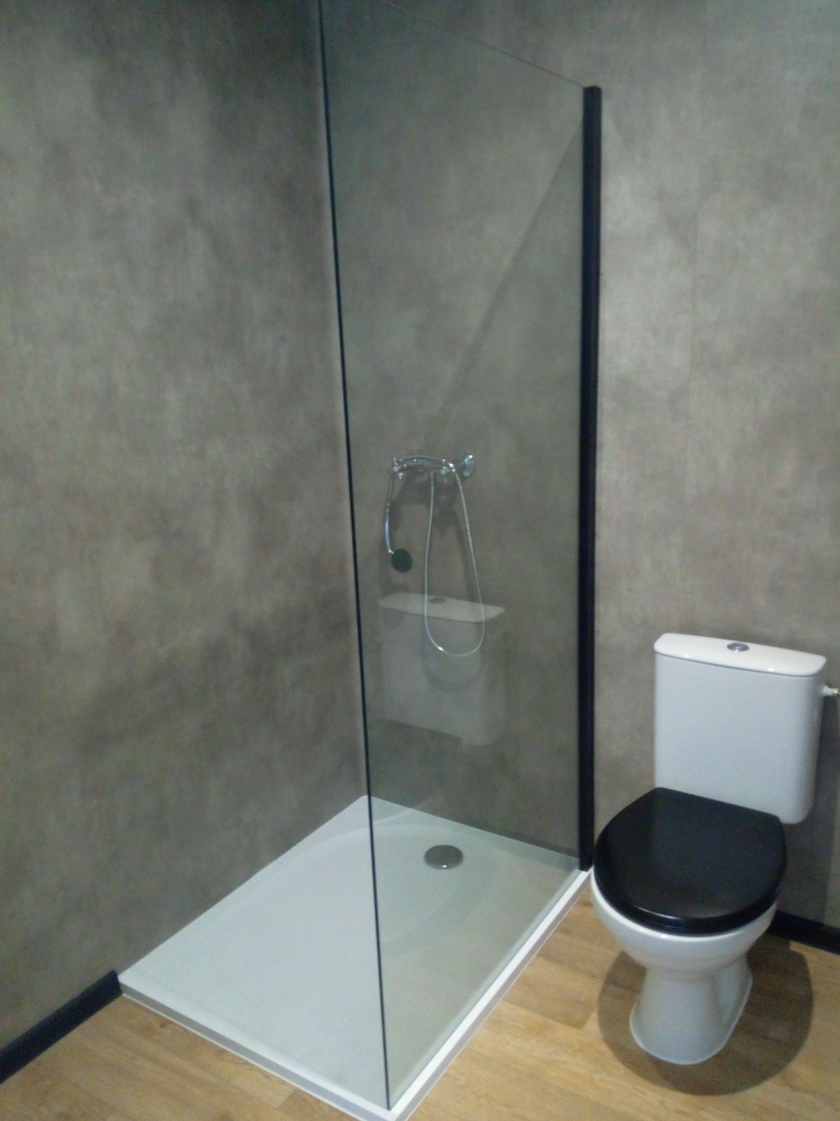 Nowoczesna kabina prysznicowa typu walk-in z czarną ramą, widoczna obok toaleta z czarną deską sedesową na tle szarej ściany z efektem betonu.