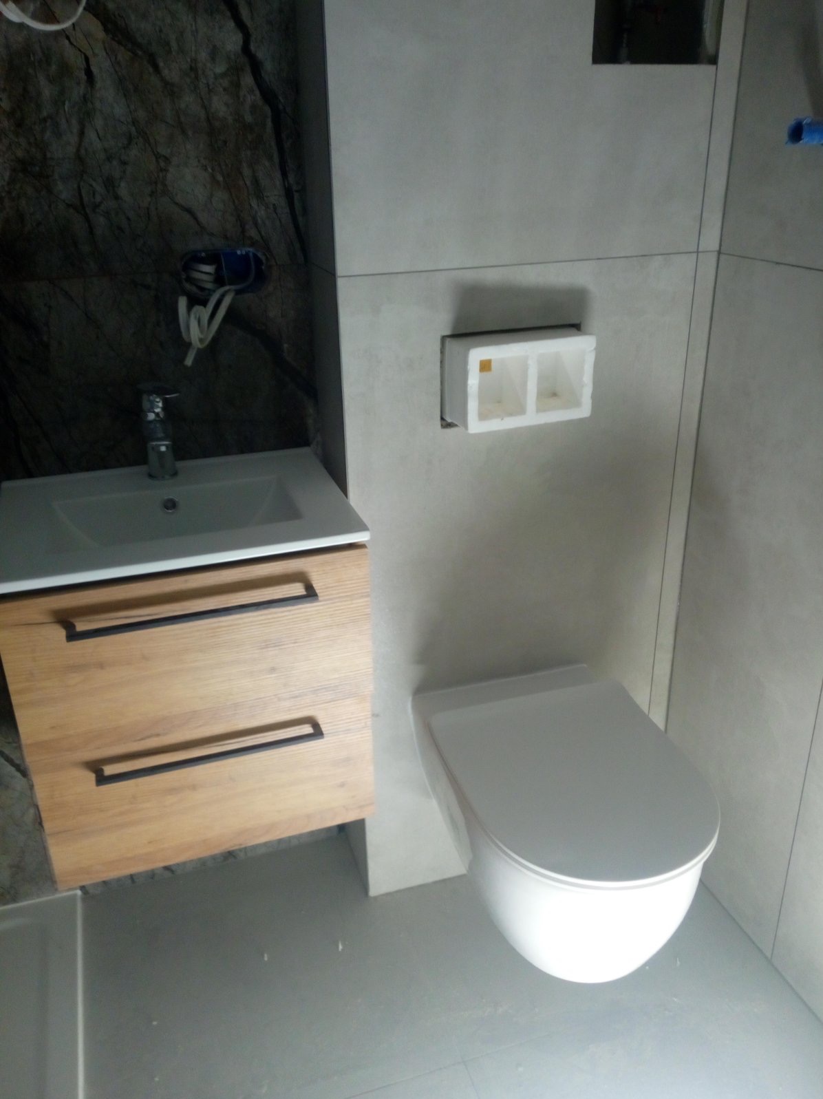 Nowoczesna łazienka z umywalką na drewnianej szafce z czarnymi uchwytami, wiszącą miską WC i szarą glazurą na ścianach, widoczne wyprowadzone kable elektryczne.