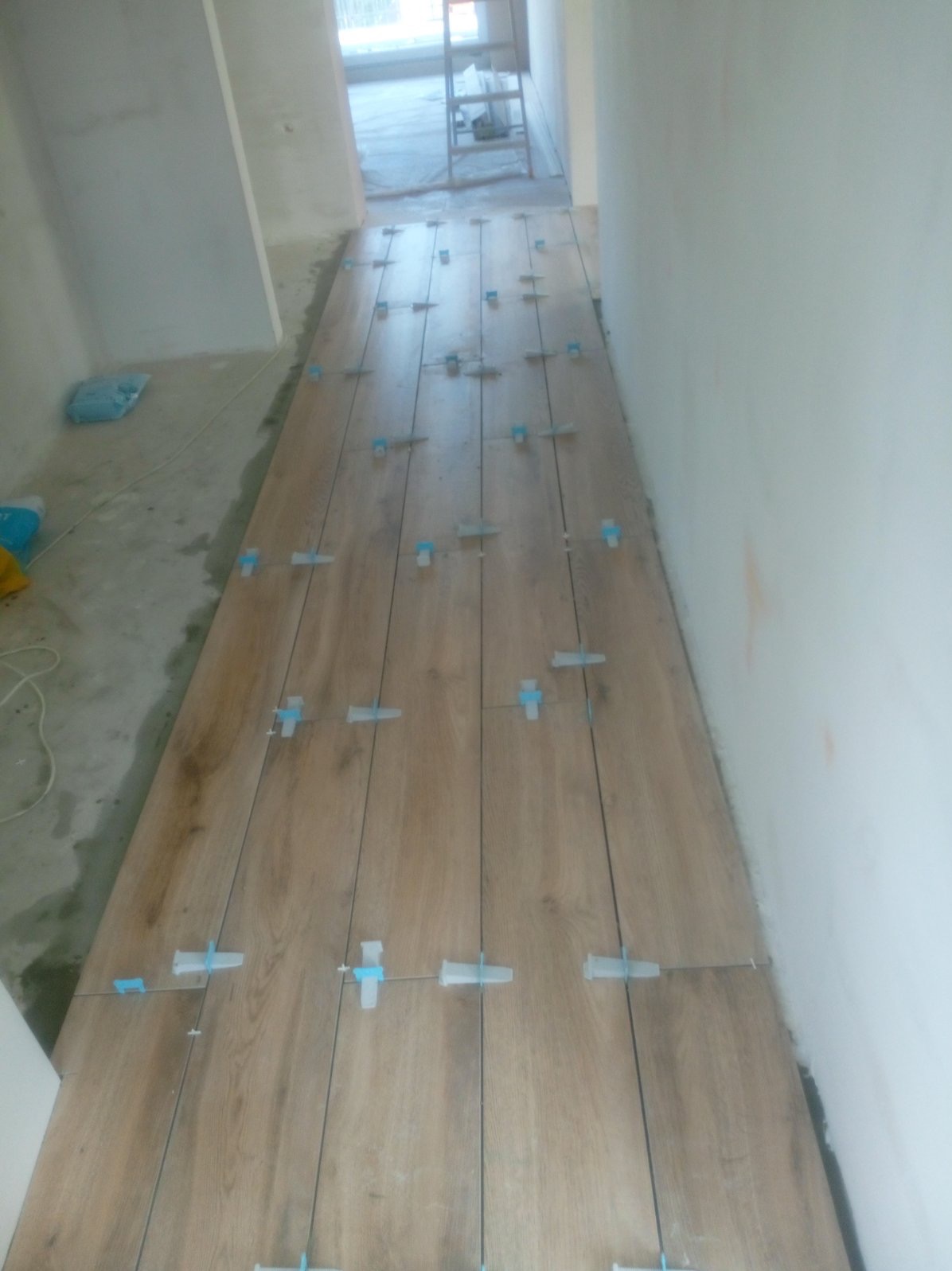 Układanie płytek imitujących drewno na podłodze korytarza, z użyciem niebieskich krzyżyków dystansowych, widok na surową ścianę i drabinę w tle.