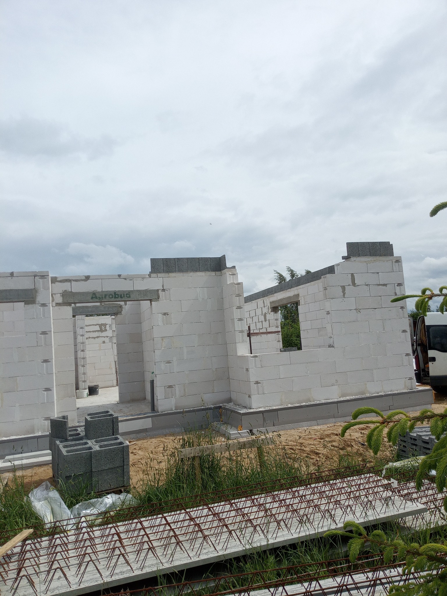 Surowy stan domu z białych bloczków betonu komórkowego, widoczne otwory okienne i drzwiowe, zbrojenie fundamentów na pierwszym planie, pochmurne niebo.
