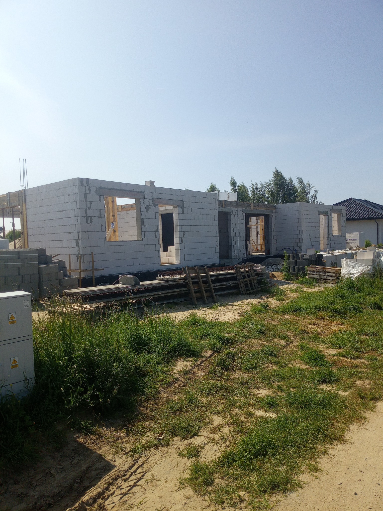 Surowy stan domu murowanego z betonu komórkowego w słoneczny dzień. Widoczne otwory okienne i drzwiowe, materiały budowlane na posesji i trawa.