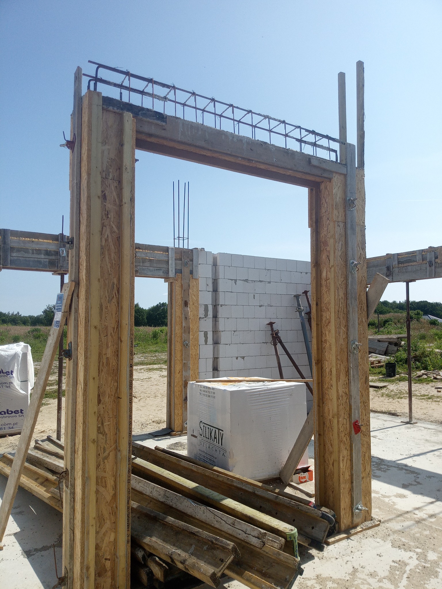 Wznoszenie nadproża z szalunkiem i zbrojeniem na budowie domu z białych bloczków silikatowych. Widoczne podpory i materiały budowlane.