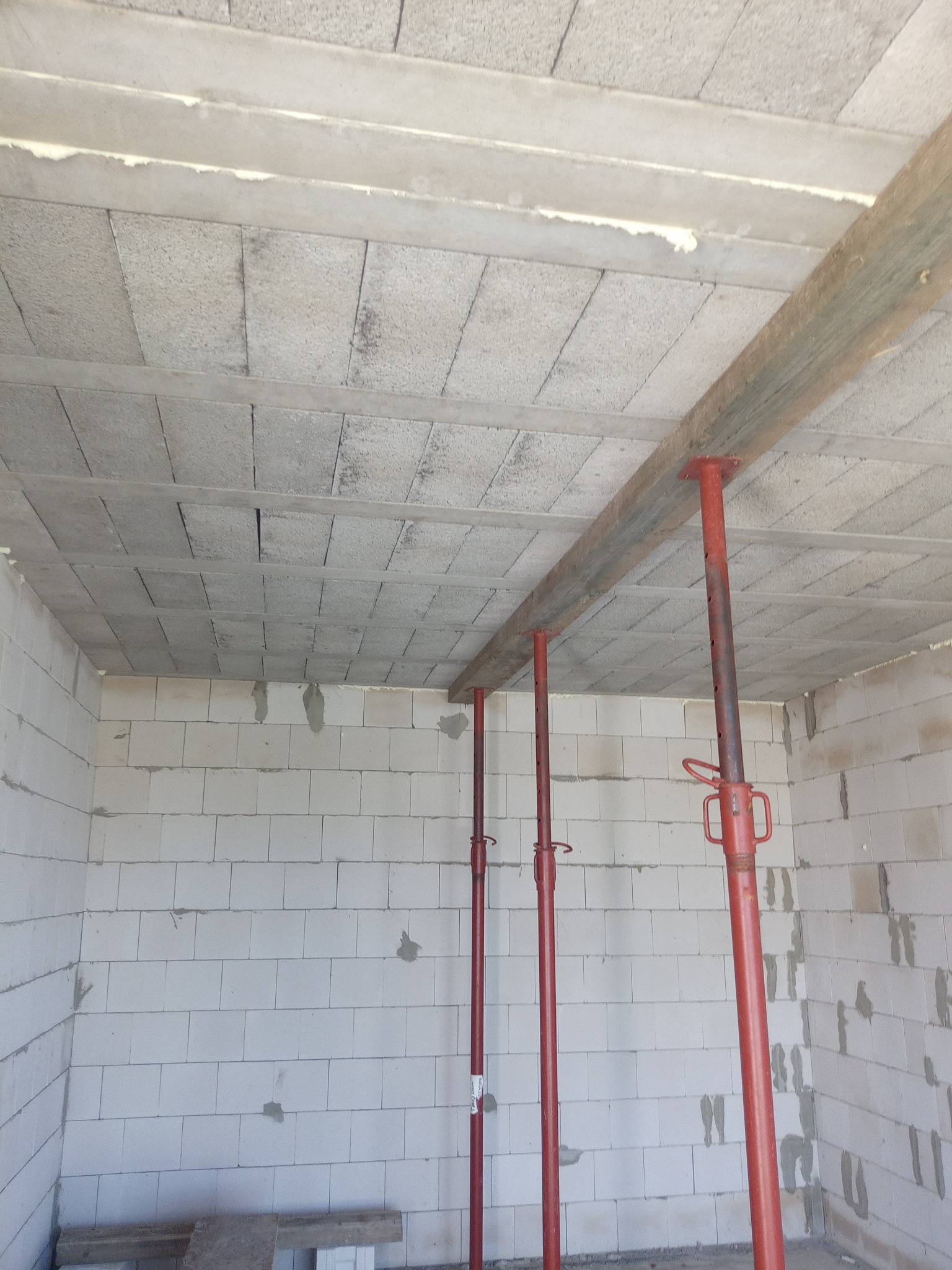 Wnętrze w trakcie budowy: ściany z białych bloczków, strop z płyt, podparty czerwonymi stemplami. Widoczne elementy konstrukcyjne i surowy stan wykończenia.